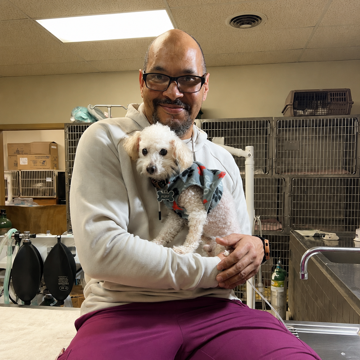 A man in glasses holding a small white dog in his arms inside a veterinary clinic or animal shelter, with cages and medical equipment in the background.