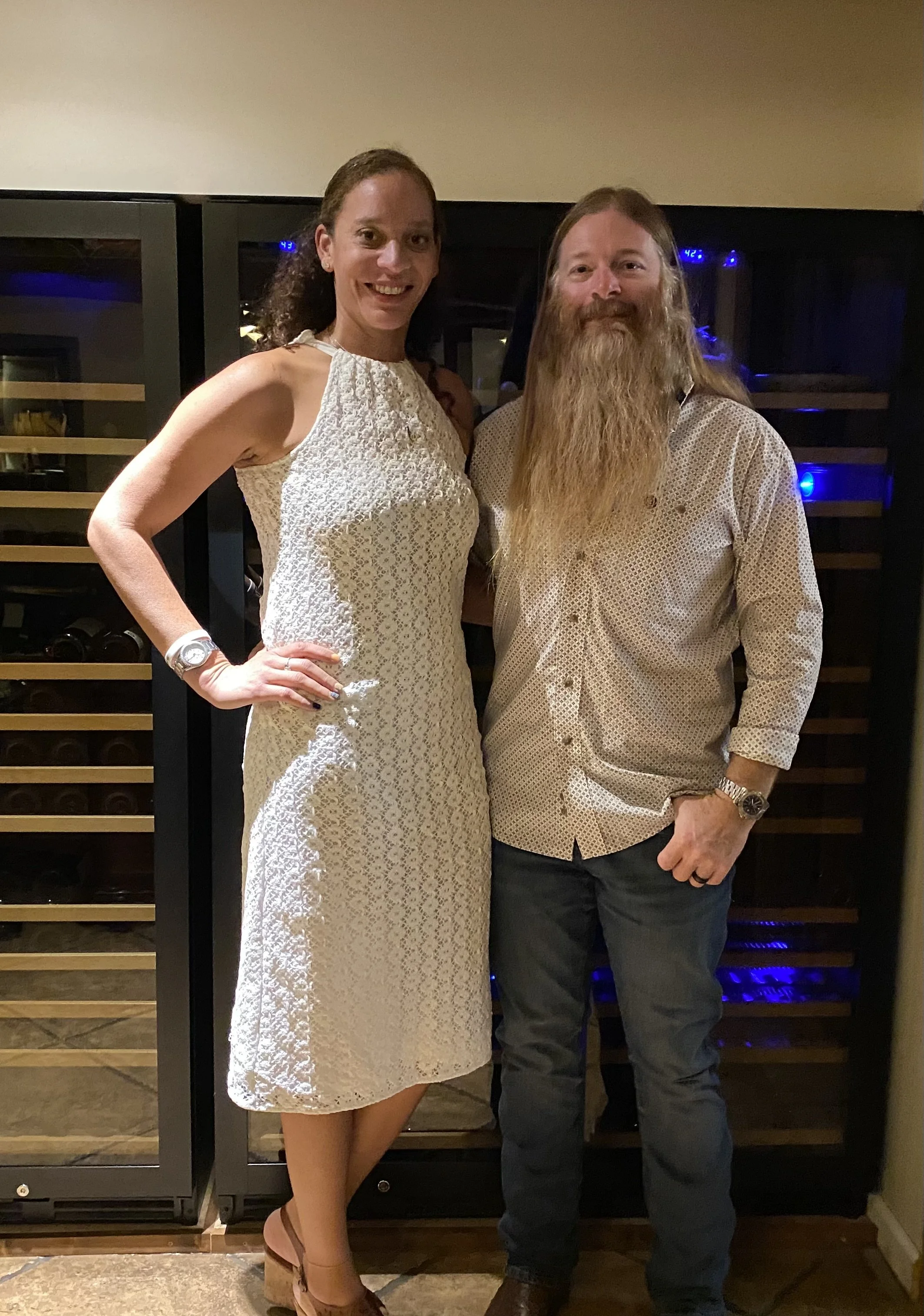 A woman in a white lace dress and tan high heels standing next to a man with long hair and beard wearing a patterned button-up shirt and jeans, posing in front of a wine storage wall in a dimly lit room.
