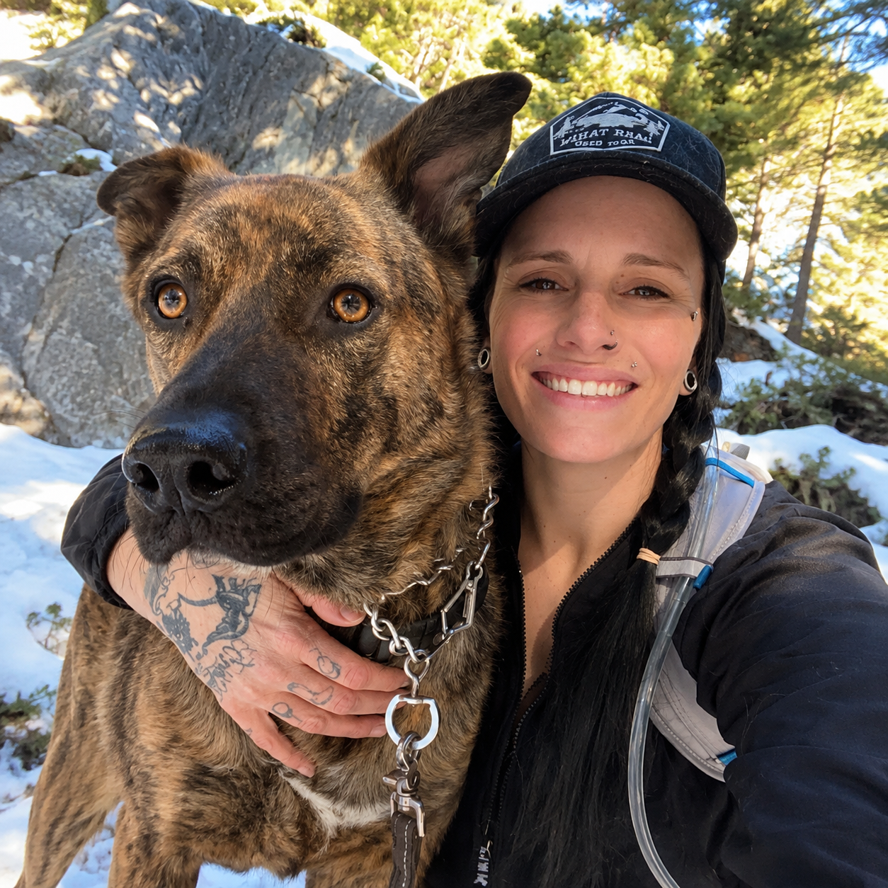 A woman in outdoor gear taking a selfie with her large brindle dog in a snowy forest setting.
