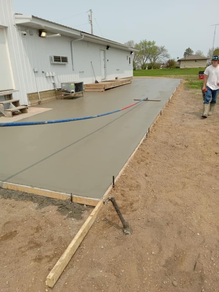 Concrete being poured and smoothed at a construction site, with a worker walking nearby. The area is bordered by wooden frames, and there is a house in the background.