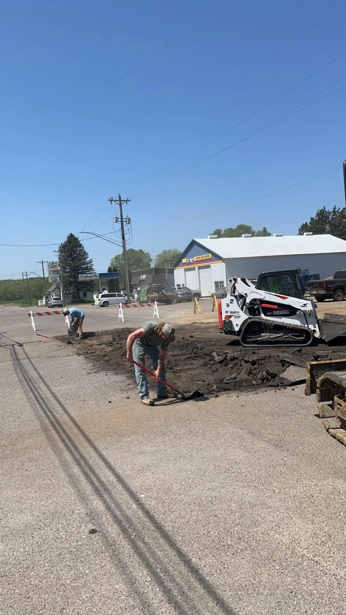 Workers are repairing the road surface with a small construction vehicle and tools, while setting up traffic barriers in a small town or commercial area.