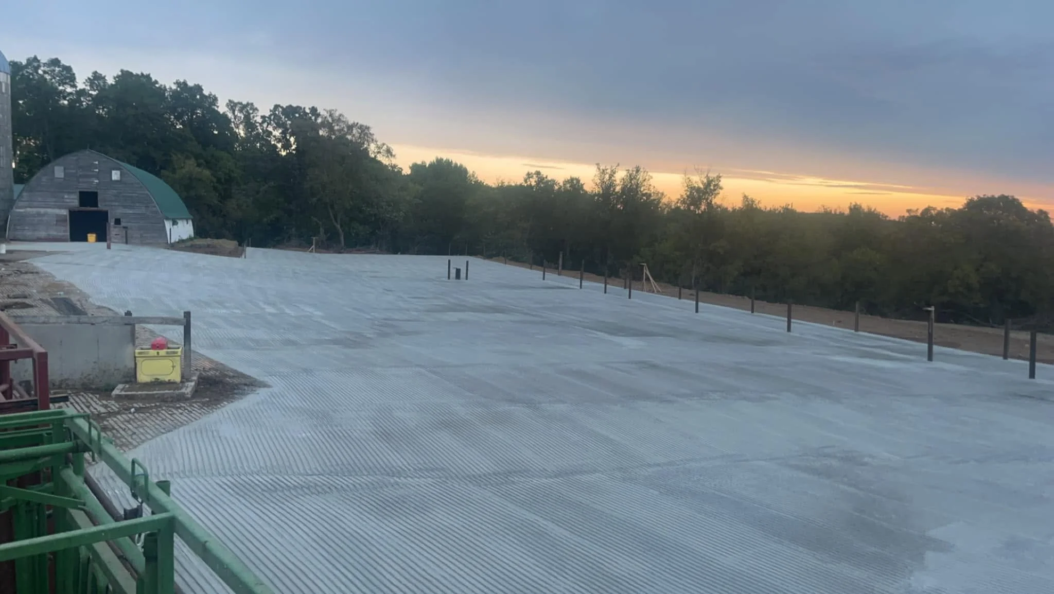 A construction site during sunset, with a partially built or covered foundation, surrounded by greenery and trees in the background.
