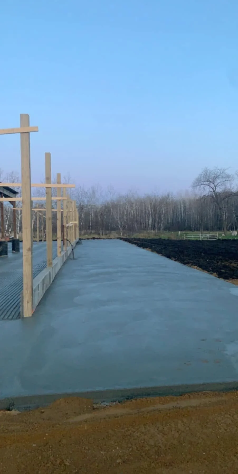 A freshly poured concrete slab foundation for a building, with wooden framing and an outdoor rural landscape in the background under a blue sky.
