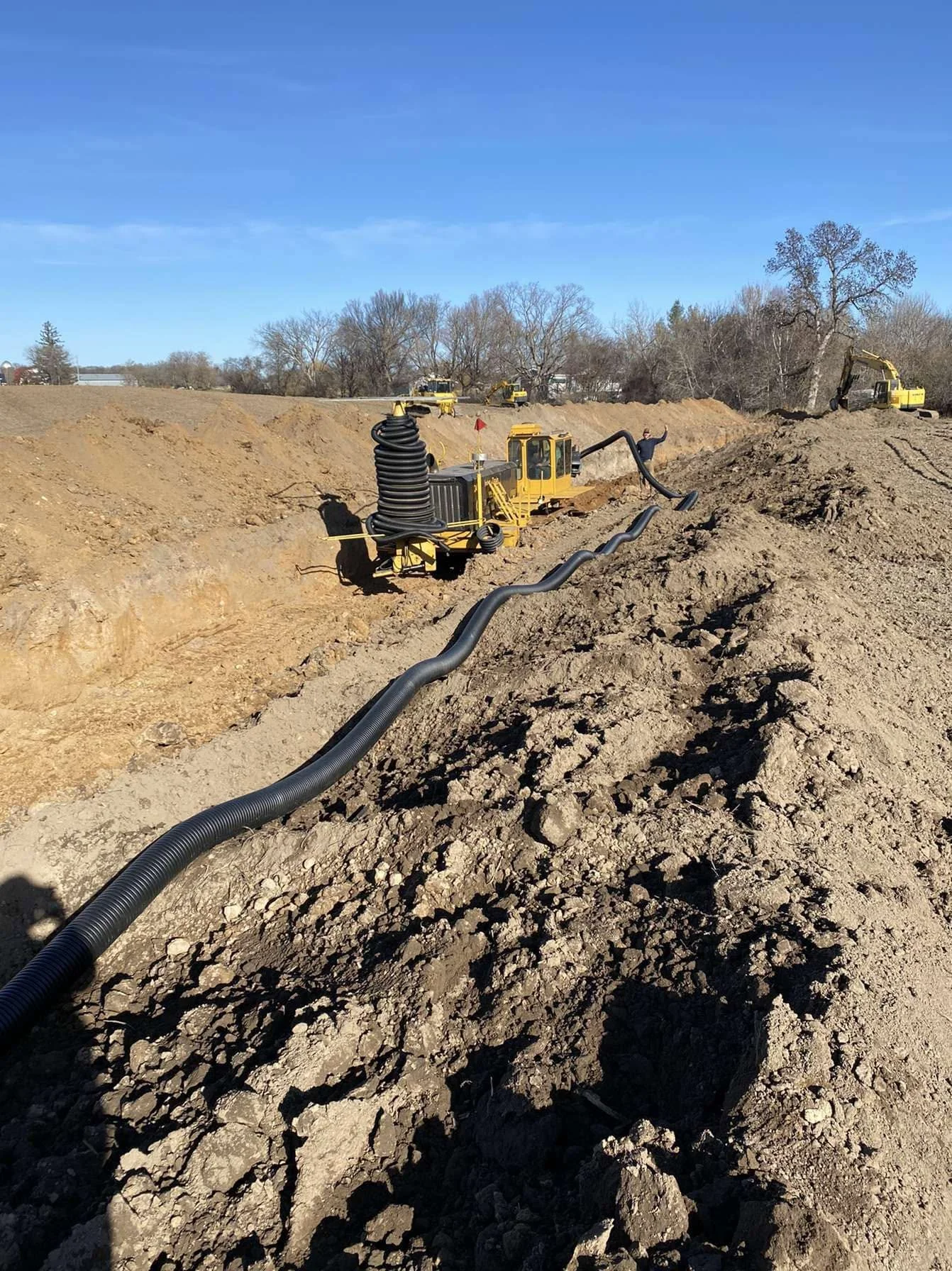 Construction site with heavy machinery, excavator, and black pipes laid out on a dirt field, with trees and a clear blue sky in the background.