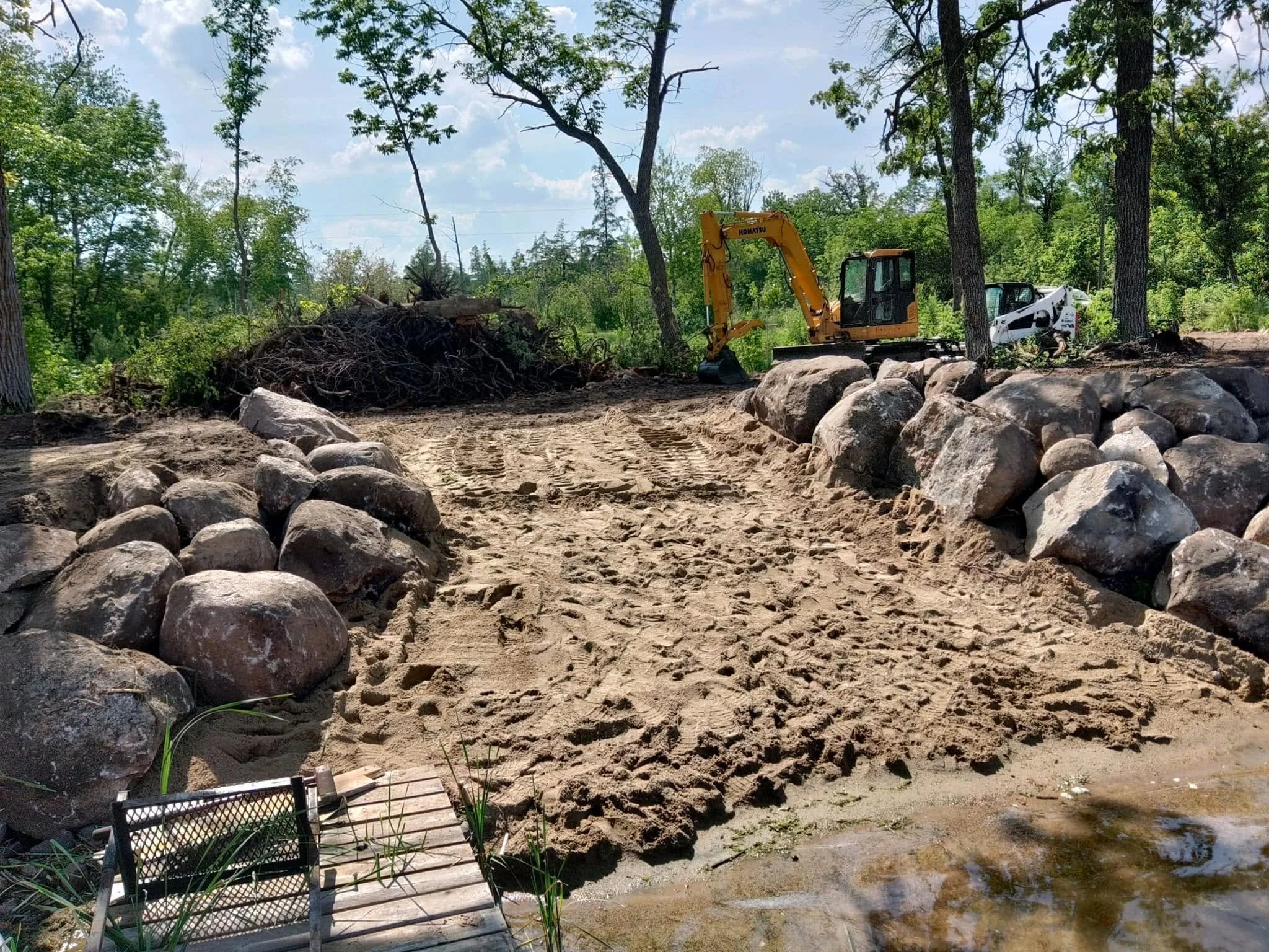 A construction scene in a wooded area with a yellow Komatsu excavator and a small white skid-steer loader. The ground is excavated with large rocks and dirt, and there are trees and a sky with clouds in the background.
