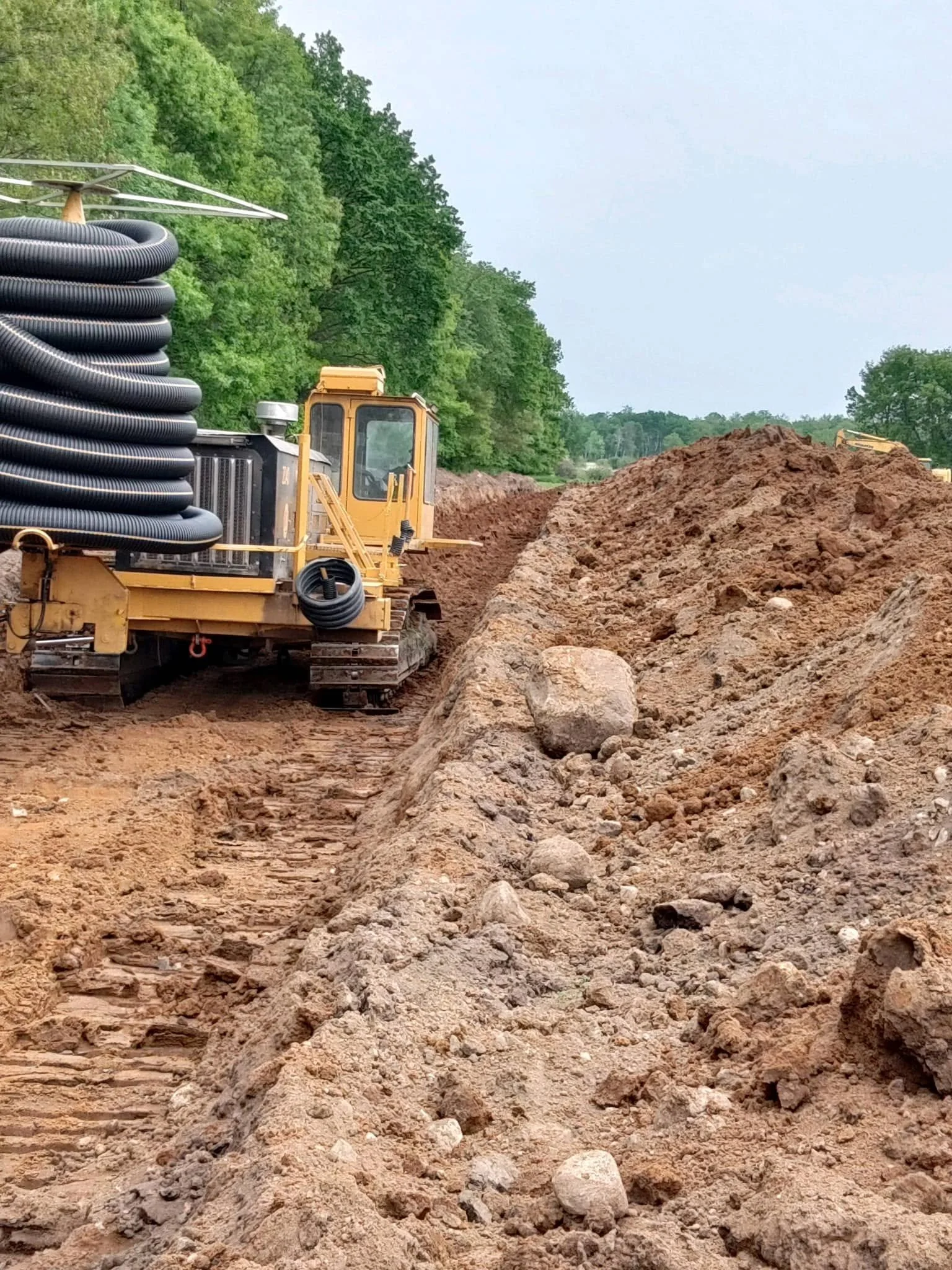Construction site with a large pile of dirt and a bulldozer. A hose is coiled on a piece of construction equipment in the foreground, and green trees are in the background.