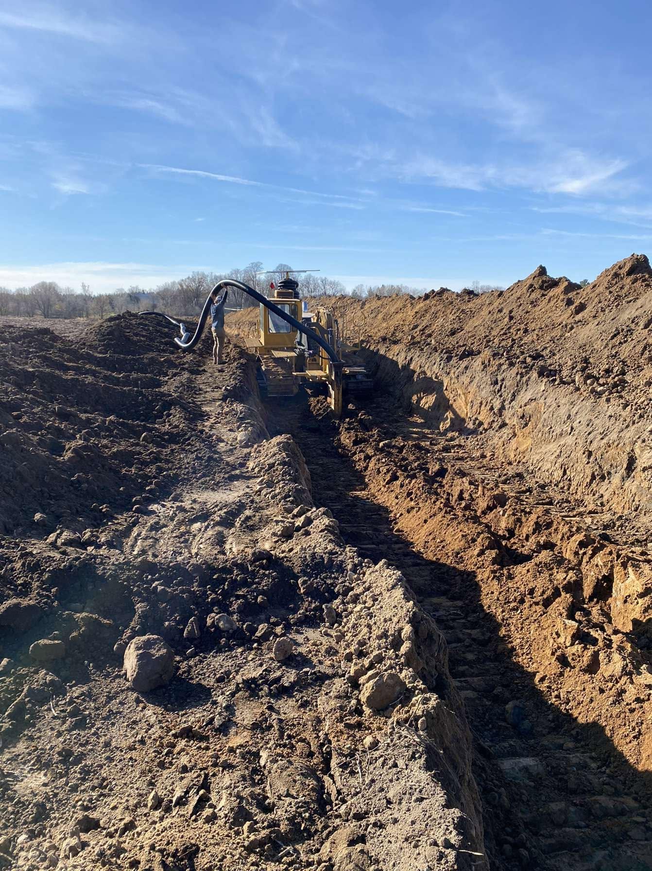 View of a construction site with heavy machinery and a large trench, with dirt piles on both sides under a clear blue sky.