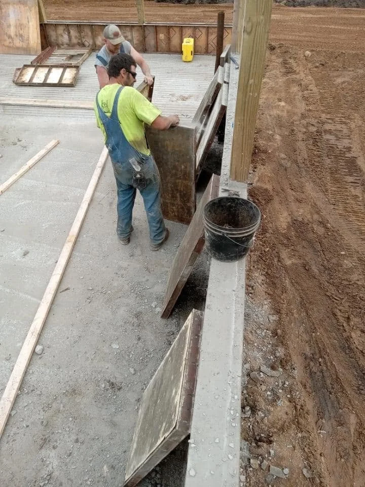 Two construction workers are installing a concrete form on a building site. One worker is holding a large wooden panel, and the other is adjusting it. The site has a partially poured concrete base, and there are tools and materials around, including 