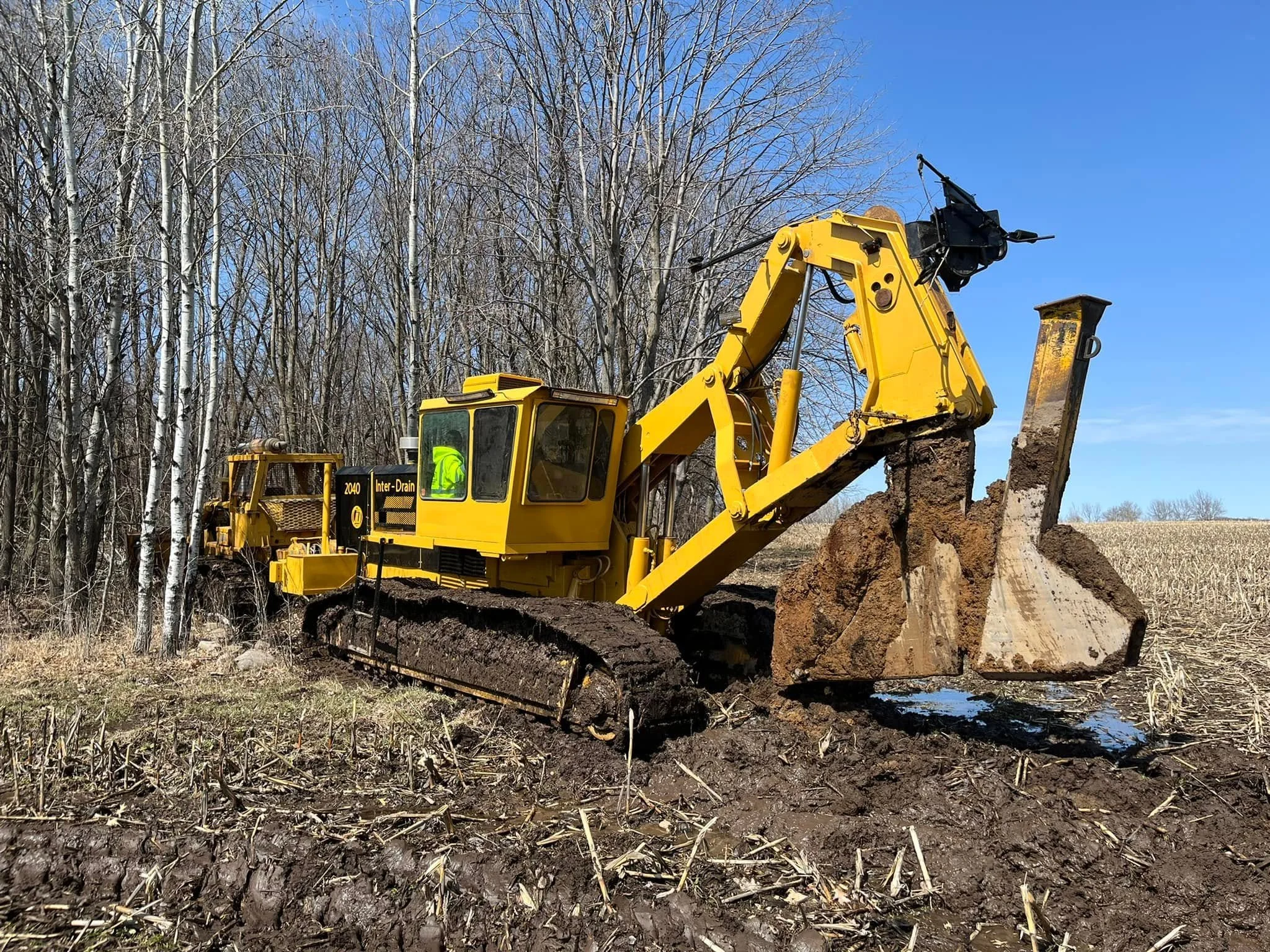 A yellow bulldozer moving soil on a construction site with leafless trees and a clear blue sky in the background.
