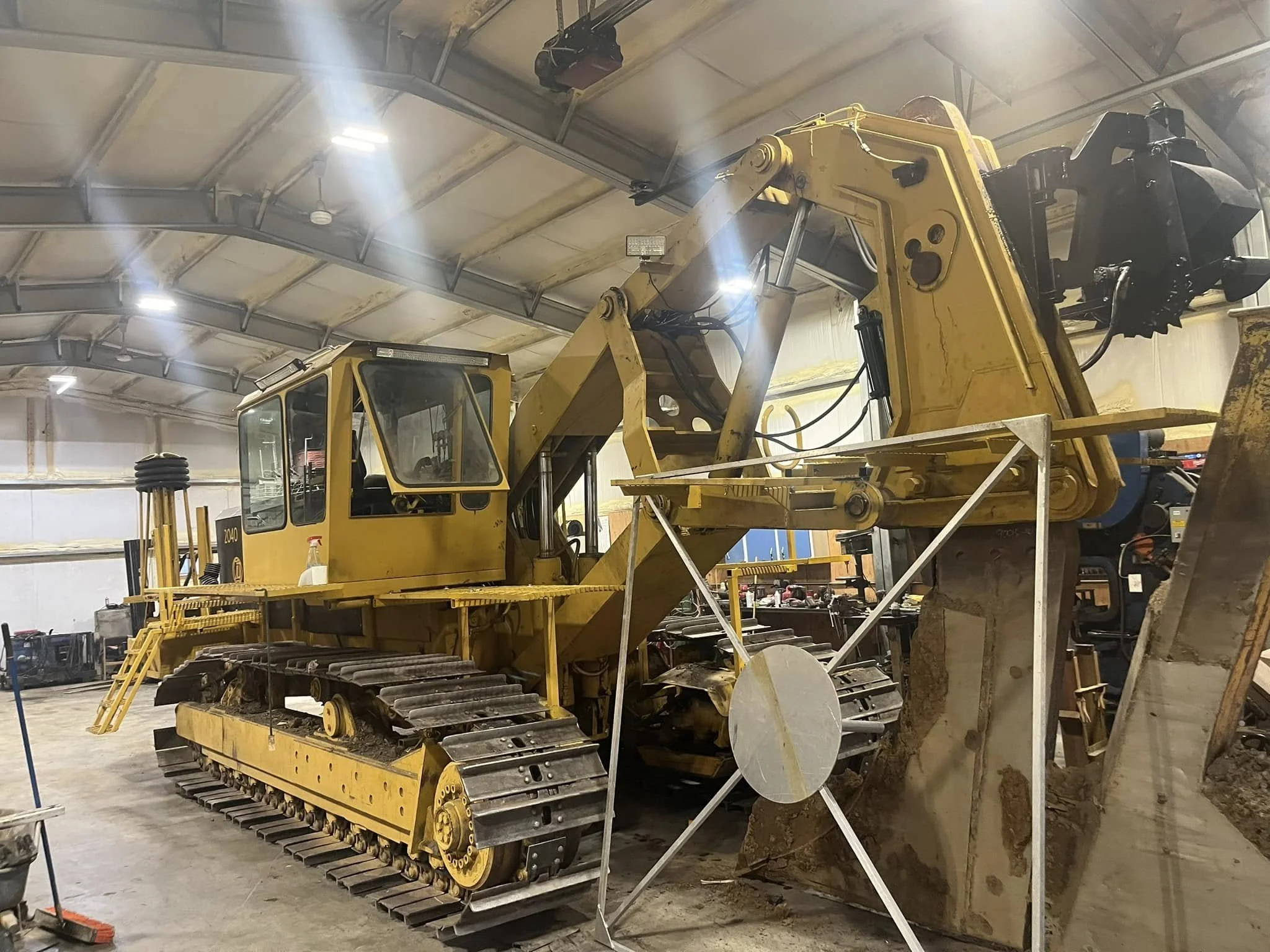 A yellow bulldozer inside a warehouse or workshop, with a large bucket at the front, tracks for movement, and a cab for the operator, surrounded by construction tools and equipment.