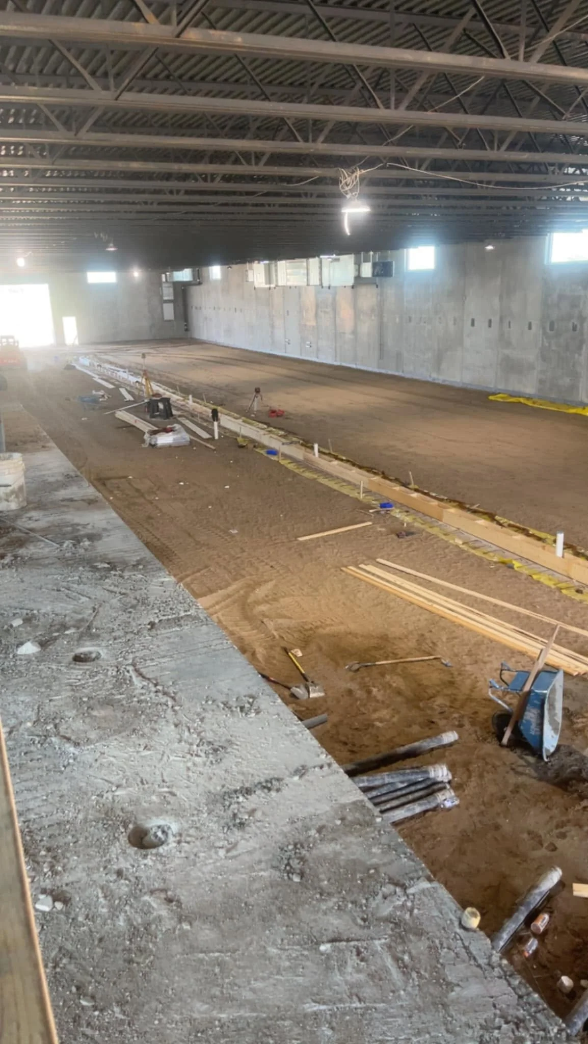 Inside an unfinished building under construction with visible metal ceiling beams, exposed concrete walls, and a dirt floor with construction materials, tools, and equipment scattered around.