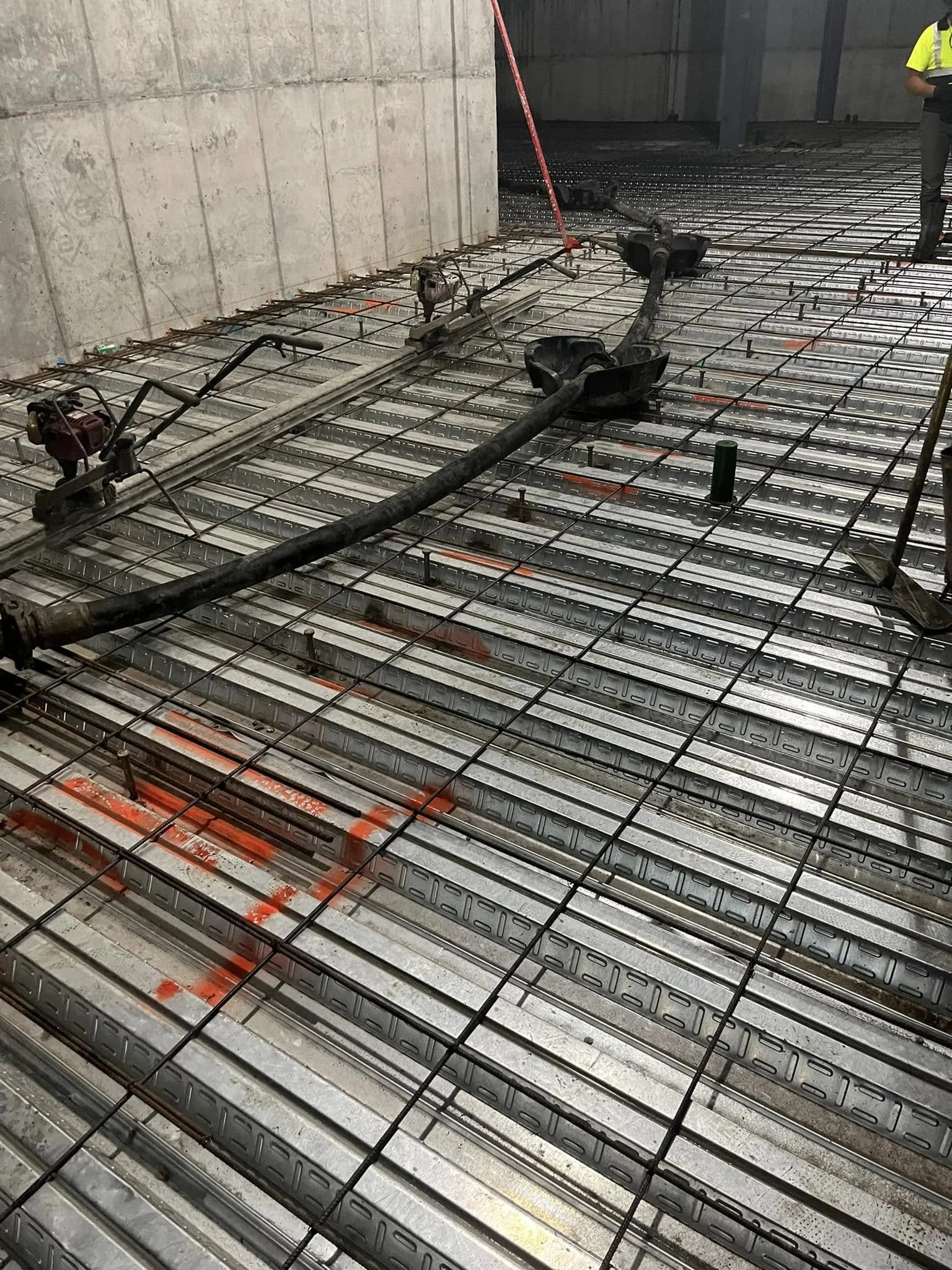 Construction site with steel reinforcement bars on the floor, pipes, and construction equipment, partially enclosed by concrete walls.