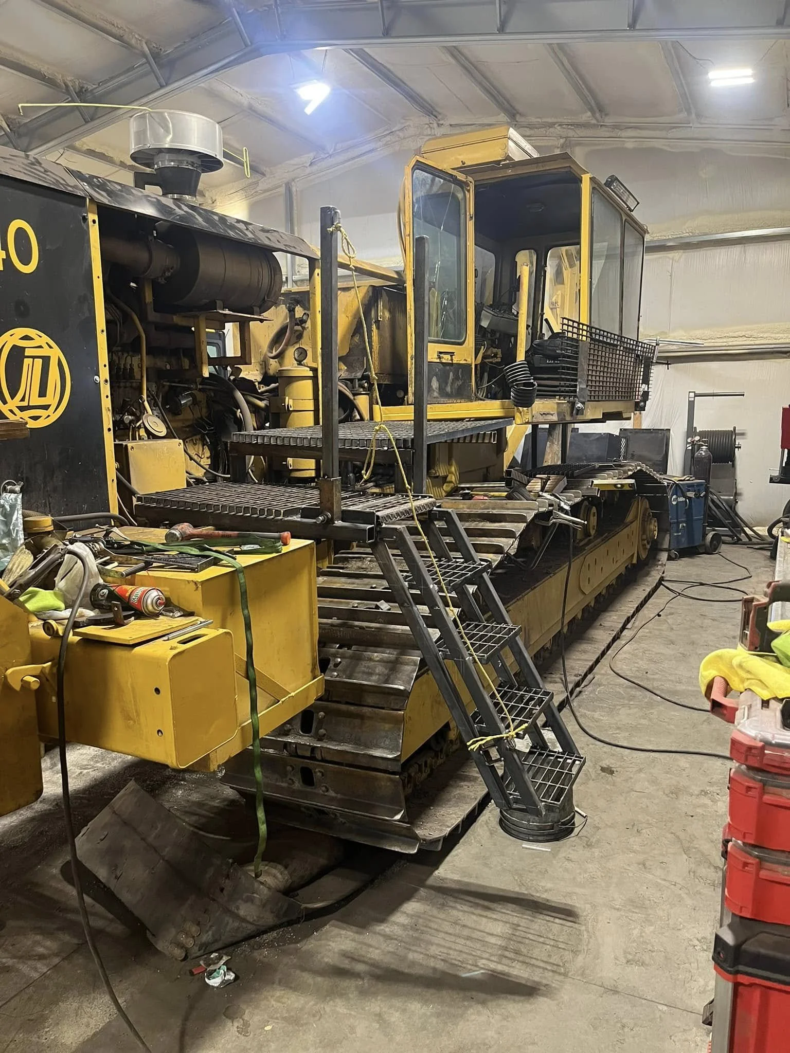 A large yellow construction bulldozer inside a workshop surrounded by tools, equipment, and barrels.