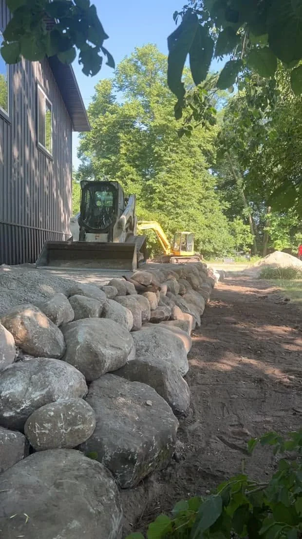 Construction site with a small bulldozer on a dirt path next to a house, with a stone retaining wall and green trees in the background.