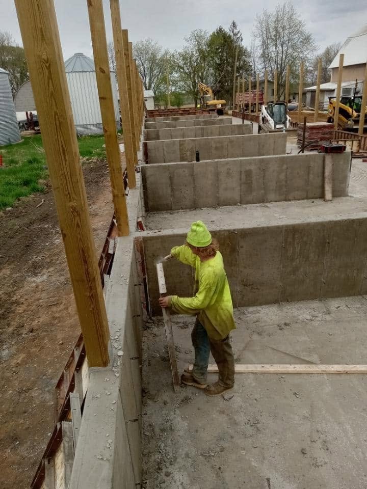 Construction worker measuring a concrete wall at a building site with wooden beams and construction equipment in the background.