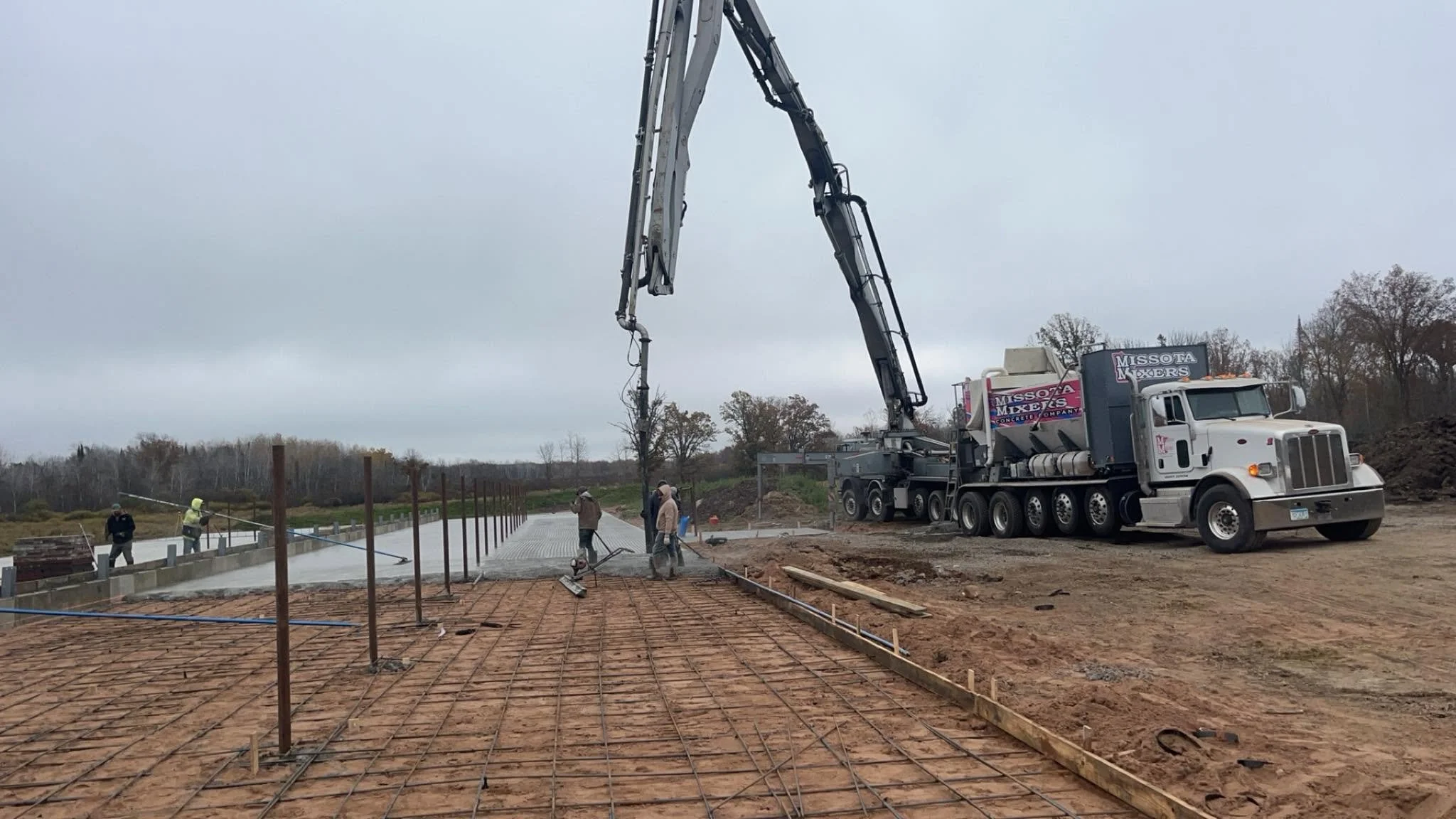 Construction site with a concrete pump truck pouring concrete onto a foundation while workers guide the flow. Rebar grid is in place for reinforcement, and wooden forms outline the edges of the foundation. Overcast sky in the background.