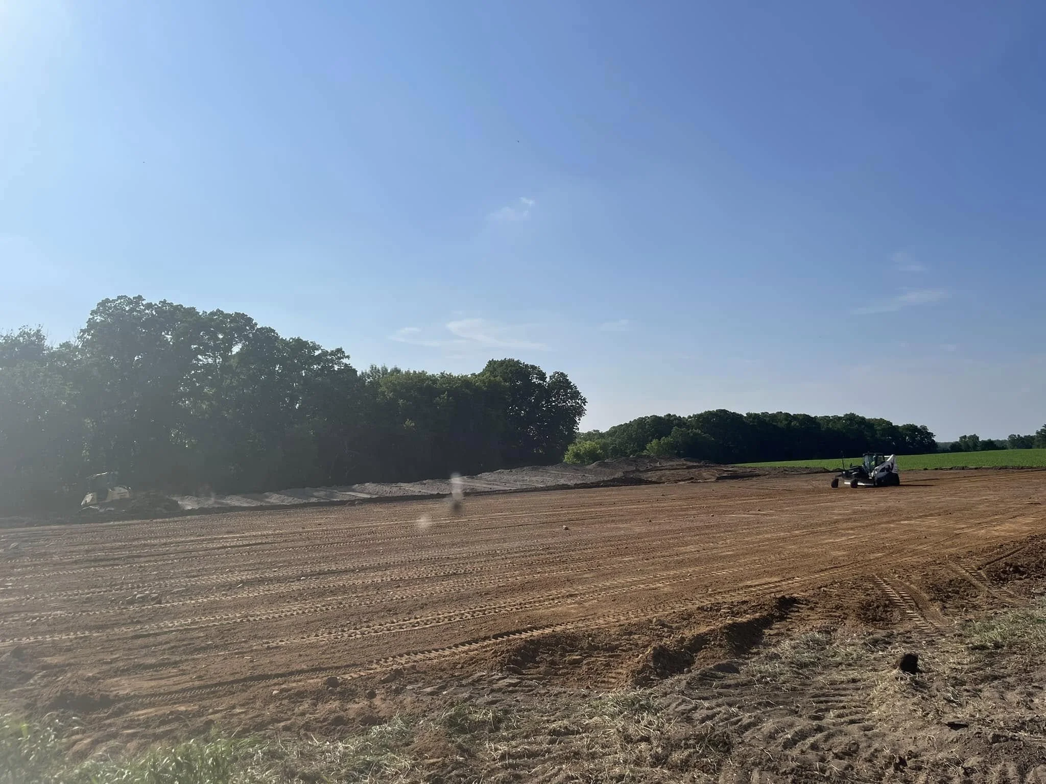 A large open field with freshly tilled soil, bordered by green trees under a clear blue sky with minimal clouds.