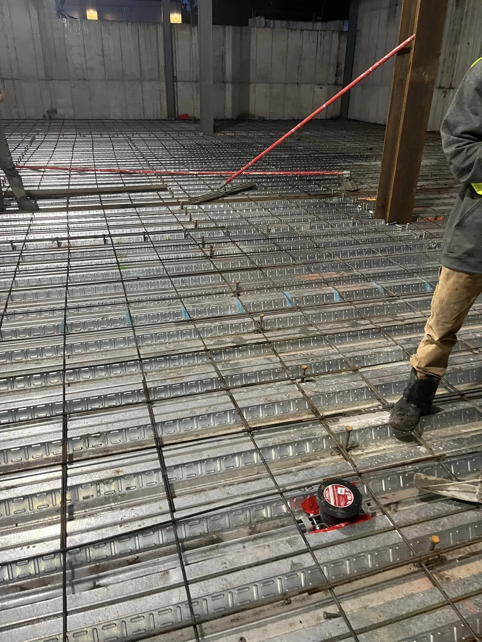 Construction site showing steel rebar grid and metal forms for concrete foundation, with workers and tools present.
