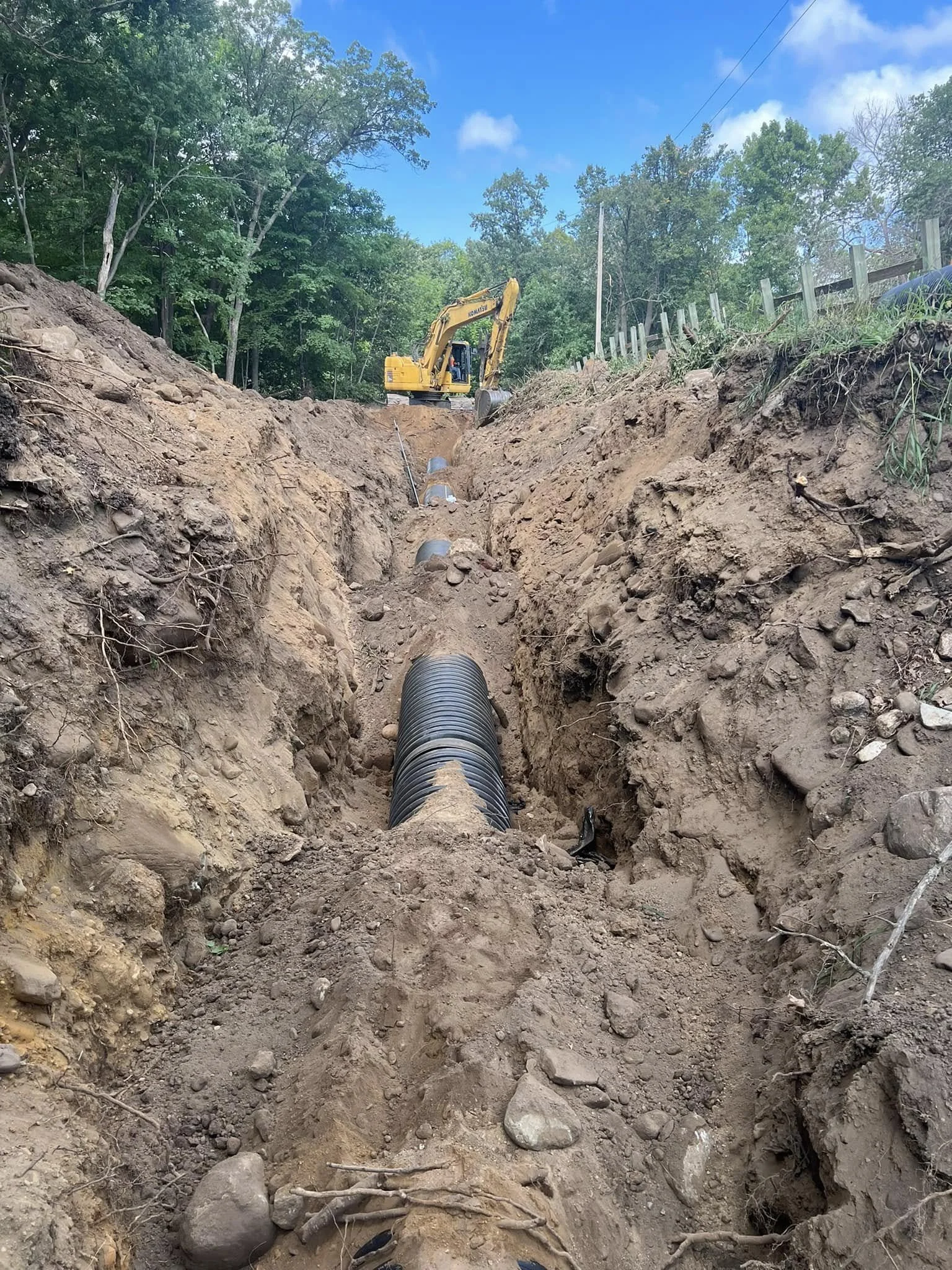 A construction site showing a large trench with a black pipe being laid underground, dirt and rocks along the sides, a yellow excavator at the top of the trench, green trees, and a partly cloudy blue sky.