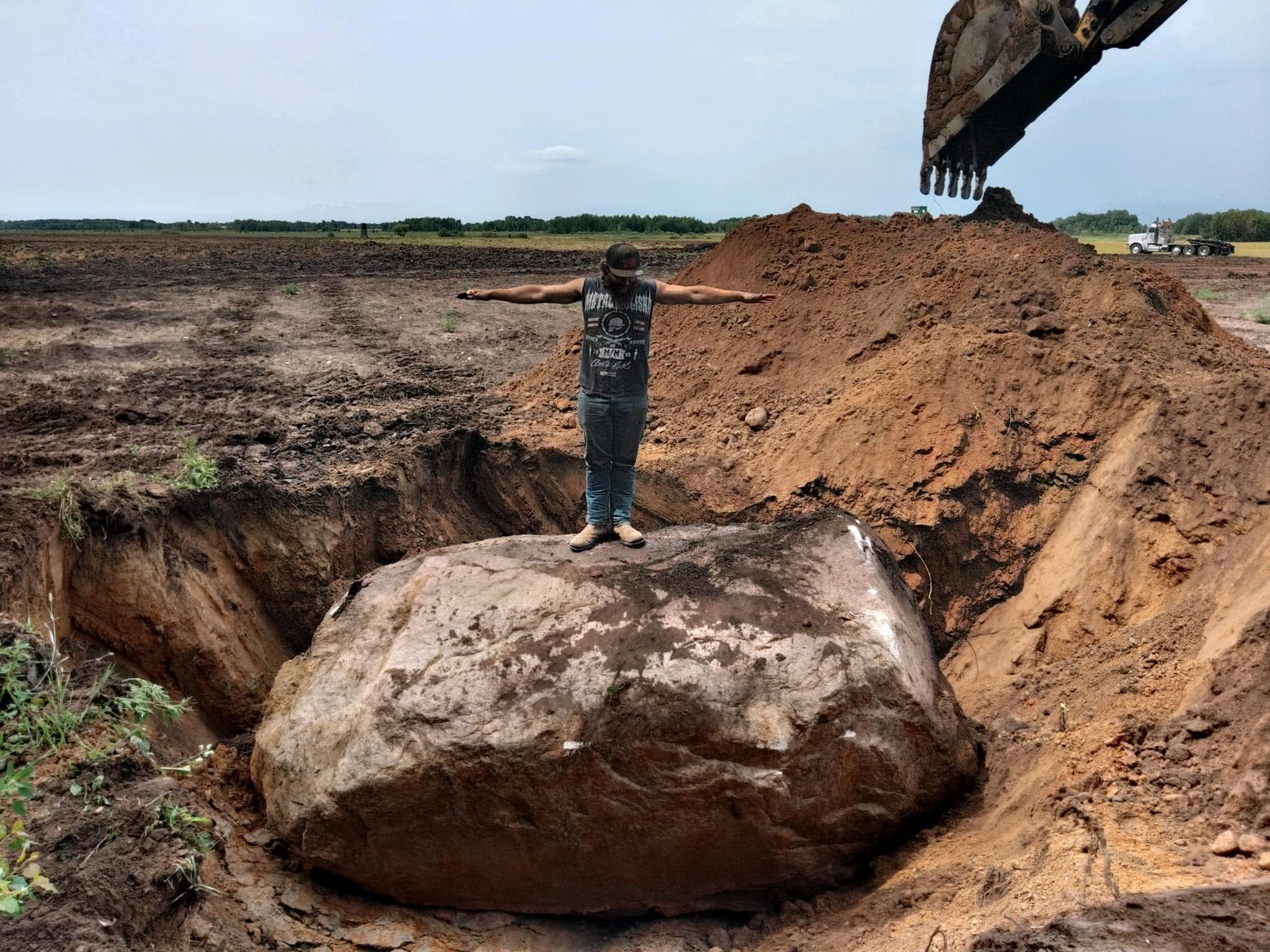 A person standing in a shallow hole with arms outstretched, next to an excavator and a large pile of dirt in an open field.