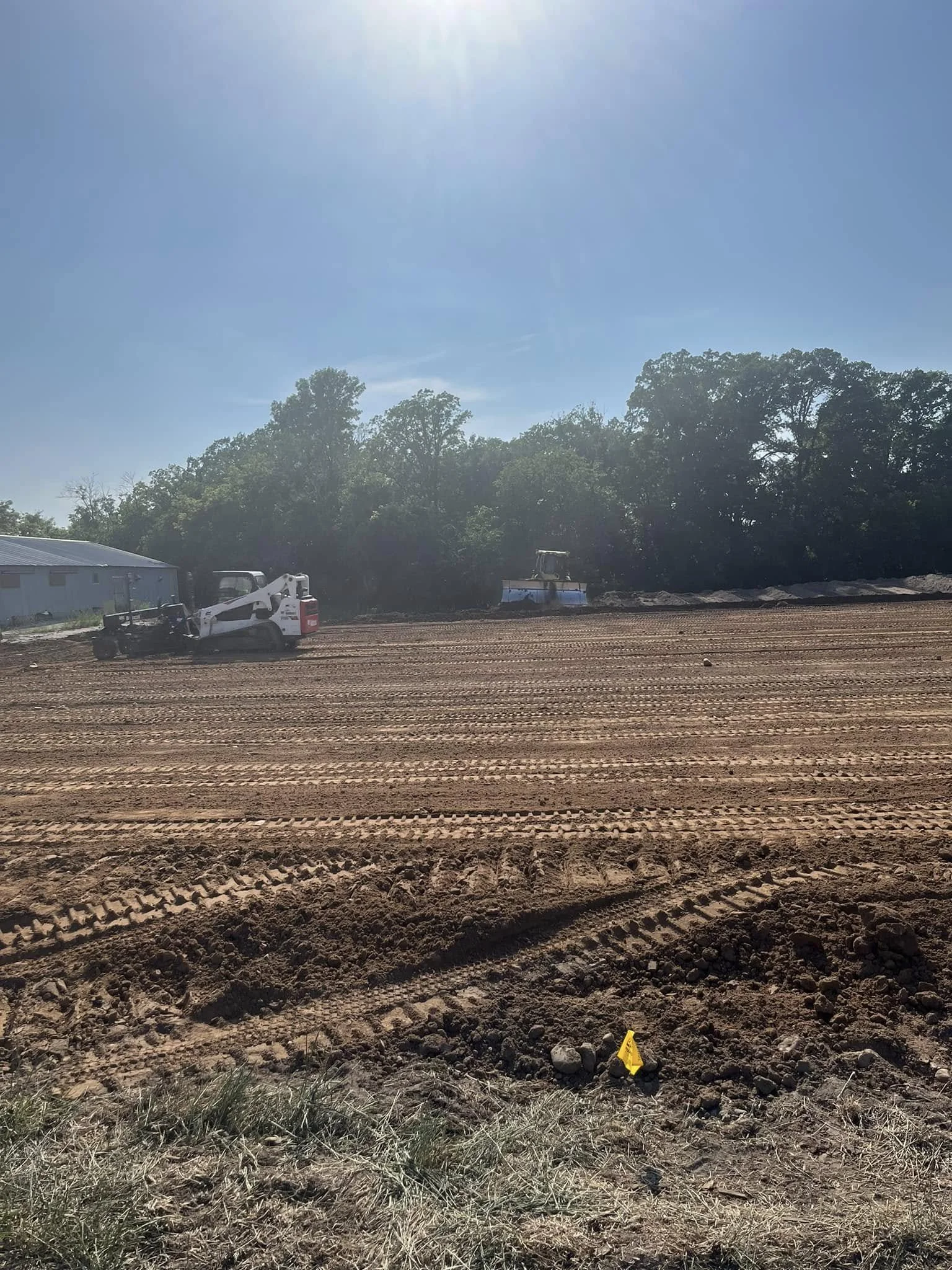 Construction site with freshly tilled soil, construction equipment, a small yellow flag, a shed, and trees under a clear blue sky.