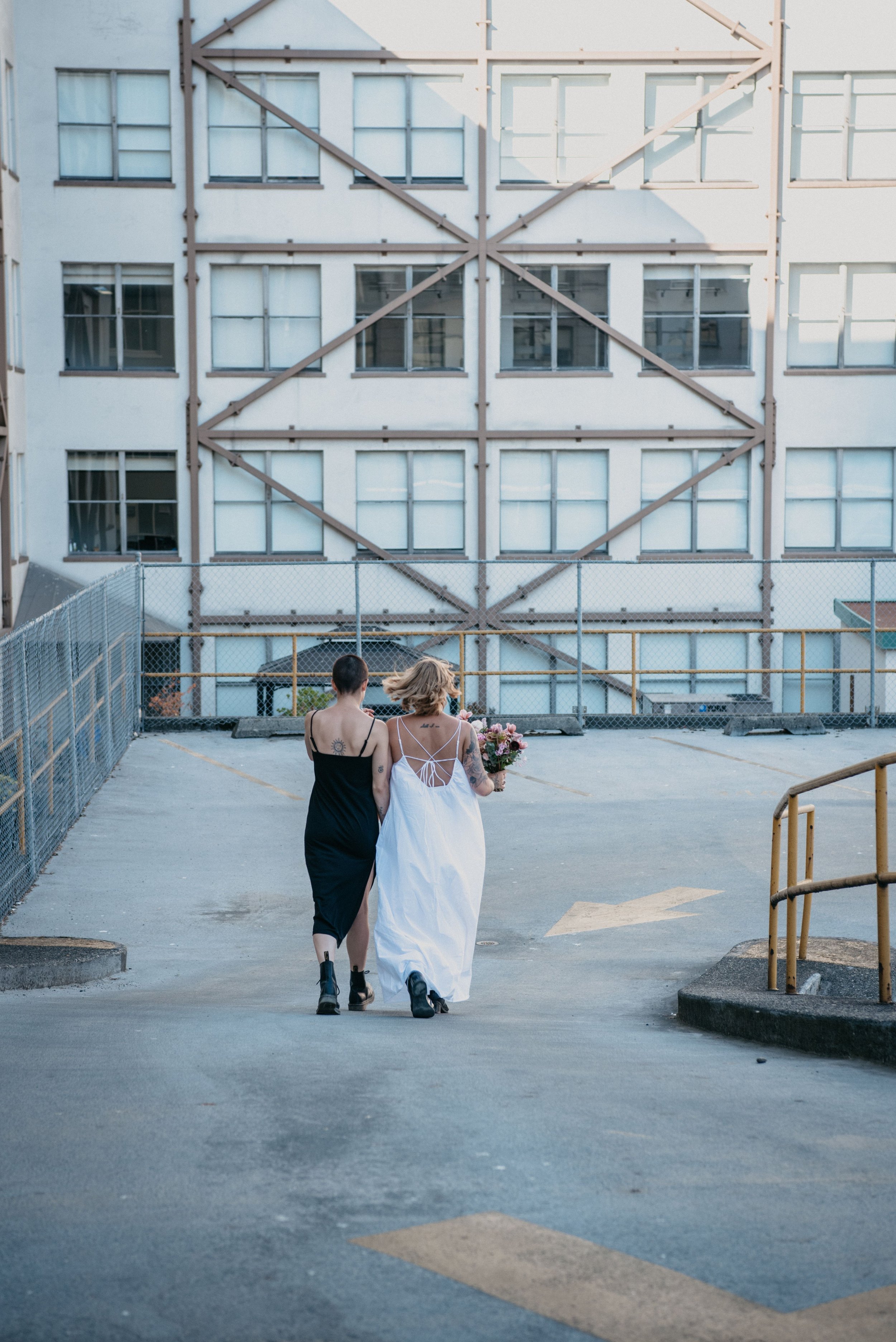 Two women with tattoos walking away from the camera in a parking lot toward a large building with multiple windows and metal framework, one in a black dress and the other in a white dress holding a bouquet of flowers.