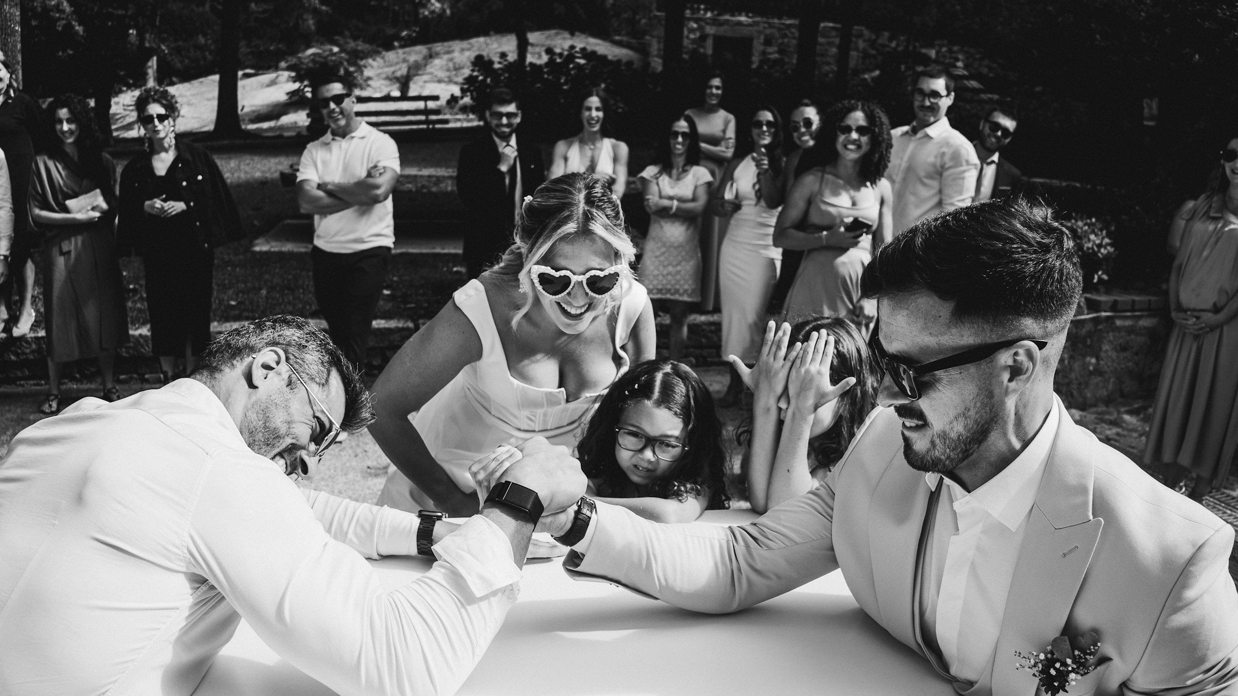 Black and white photo of a group of people gathering at a outdoor wedding, with two men in the foreground arm wrestling, woman with sunglasses laughing watching them, and others standing and smiling in the background.