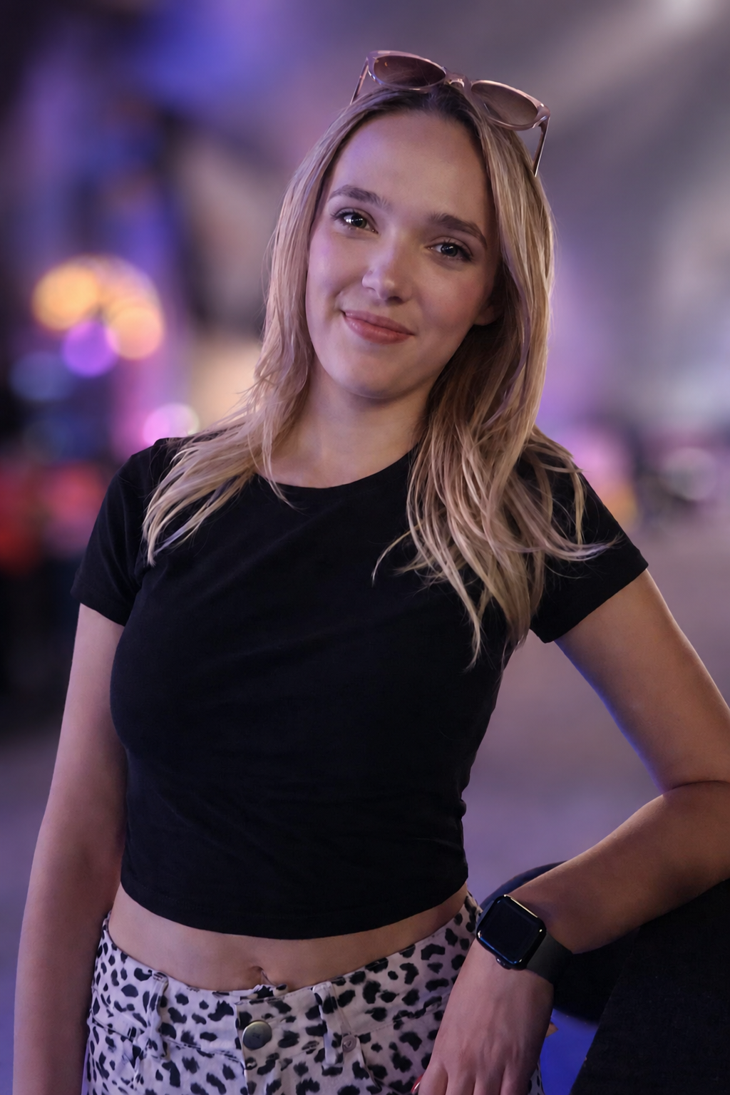 A young woman with blonde hair, wearing sunglasses on her head, a black crop top, leopard print shorts, and a smartwatch on her left wrist, standing outdoors at night with colorful lights in the blurred background.