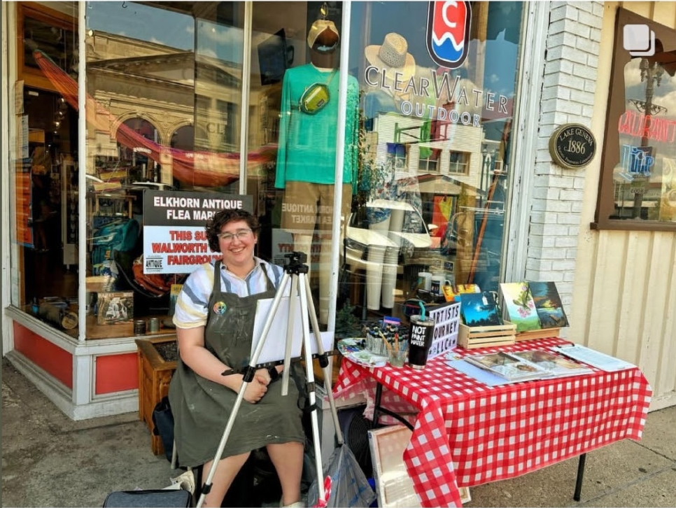 A woman sitting outside a store selling art and craft items, with a display table covered with a red and white checkered tablecloth.