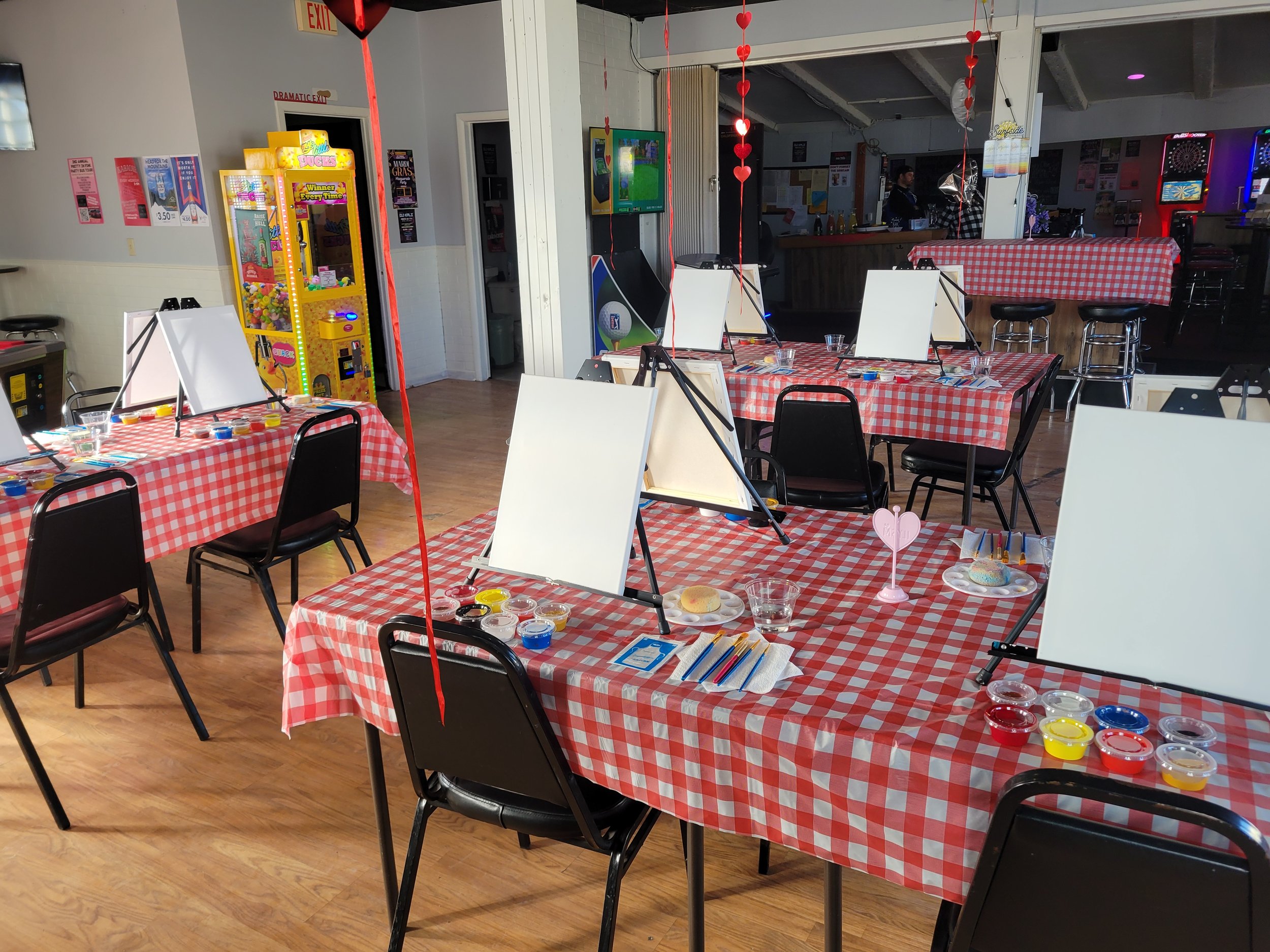 A room with red and white checkered tablecloths on tables, some with painting supplies such as water paints, brushes, and easels. Red paper hearts hang from ceiling decorations. There is a dartboard on the wall and arcade machines in the background.
