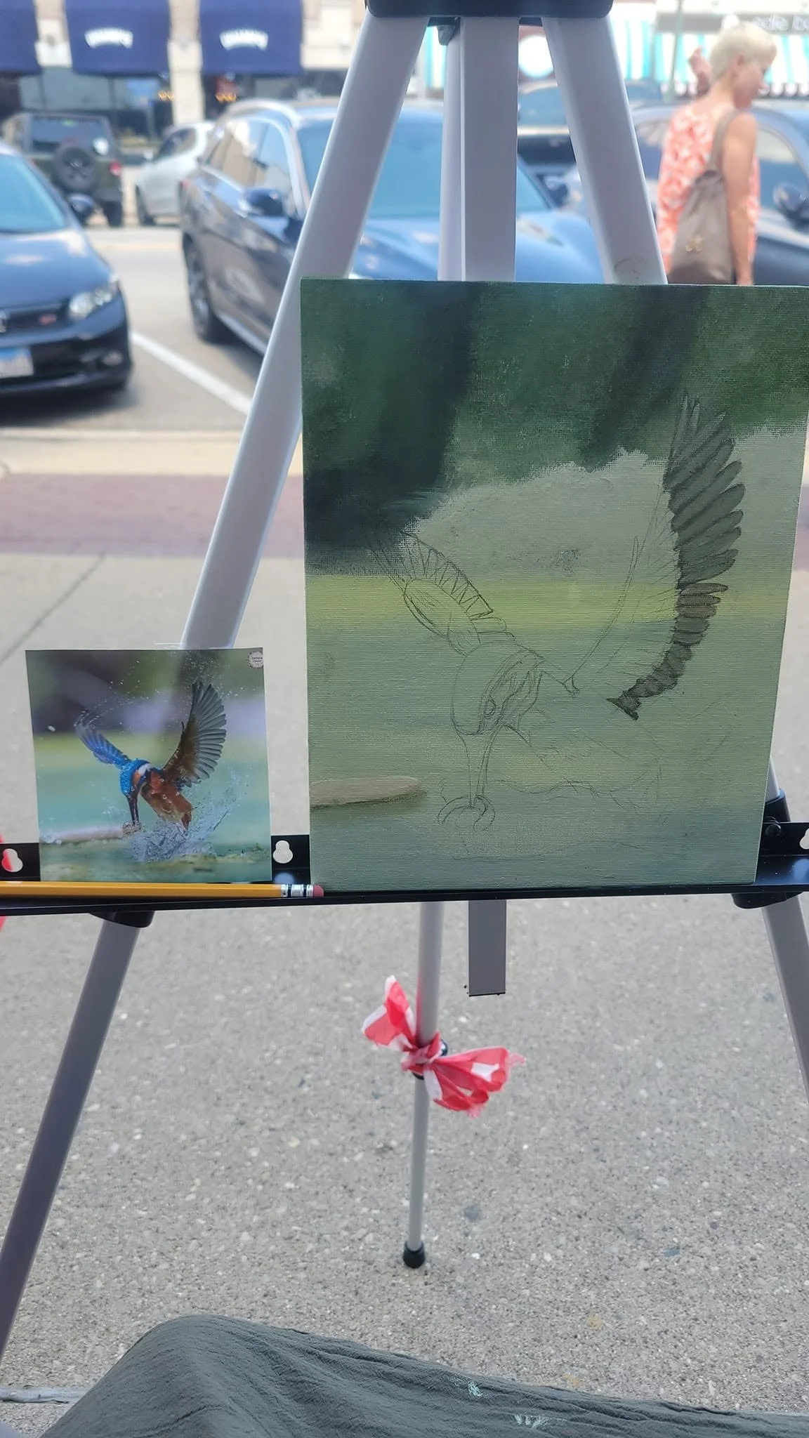 A sketch of a flying kingfisher bird on a canvas with a reference photo of a kingfisher in water, placed on an easel outdoors, with cars and two women in the background.