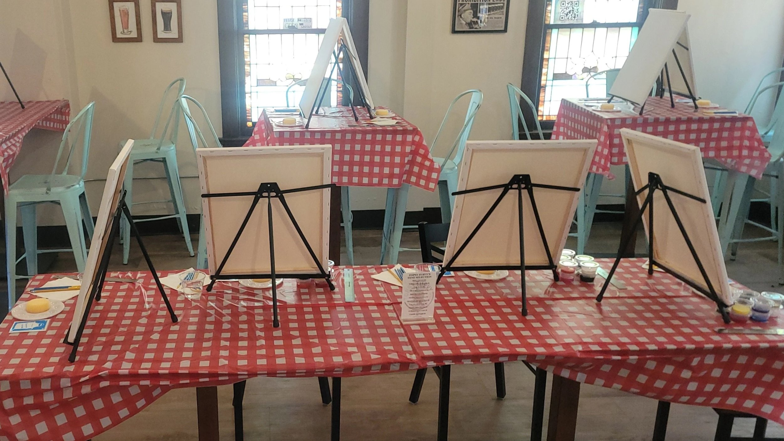 Empty restaurant table with red checkered tablecloths, set up for painting with easels, brushes, and paint cups, inside a cozy dining area with windows and art on the walls.