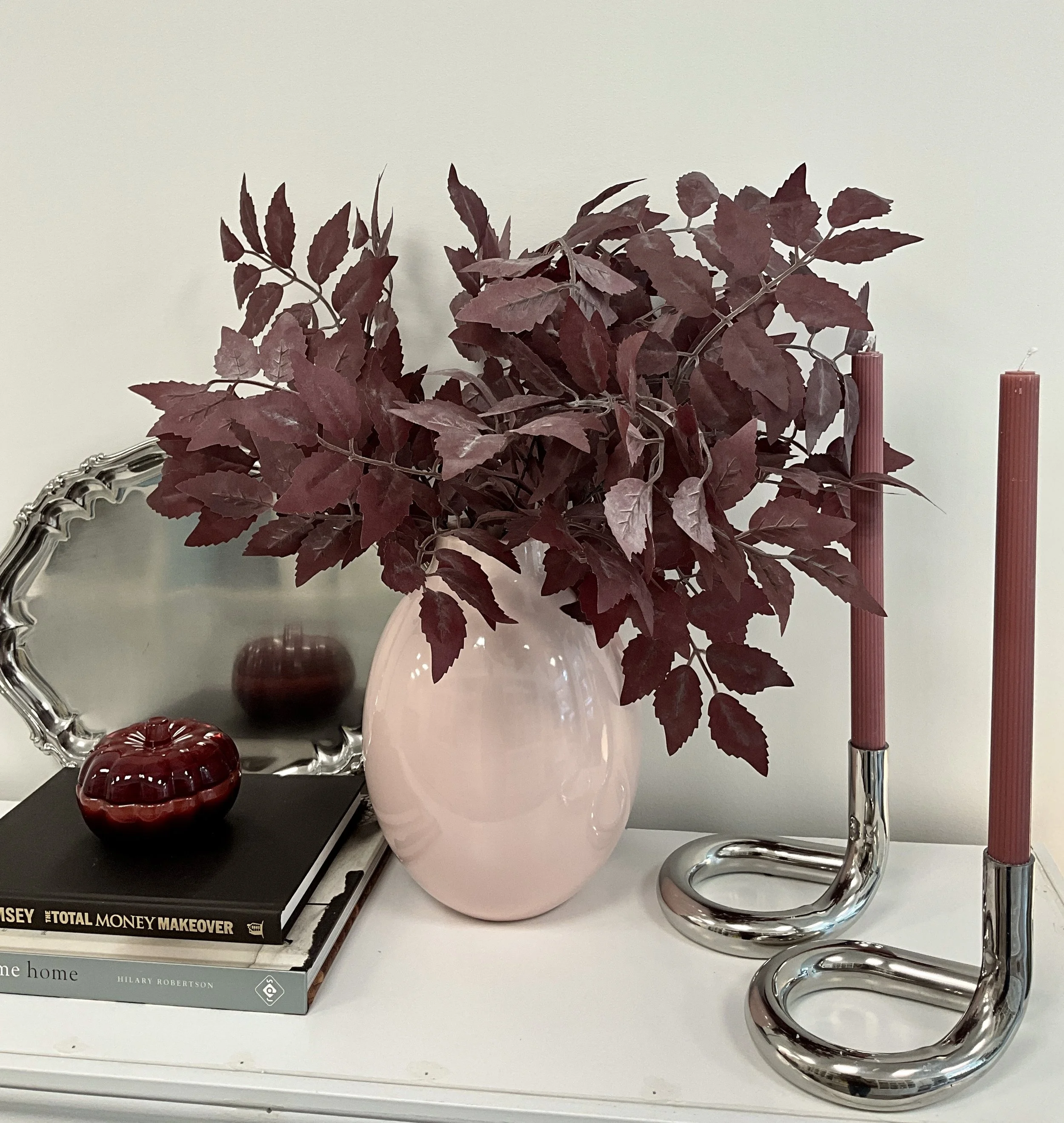Decorative display on a white shelf includes a large pink vase with burgundy leaves, two red candles in silver holders, a round red decorative object, and a stack of books including 'The Total Money Makeover' and 'Home' by Hilary Robertson.