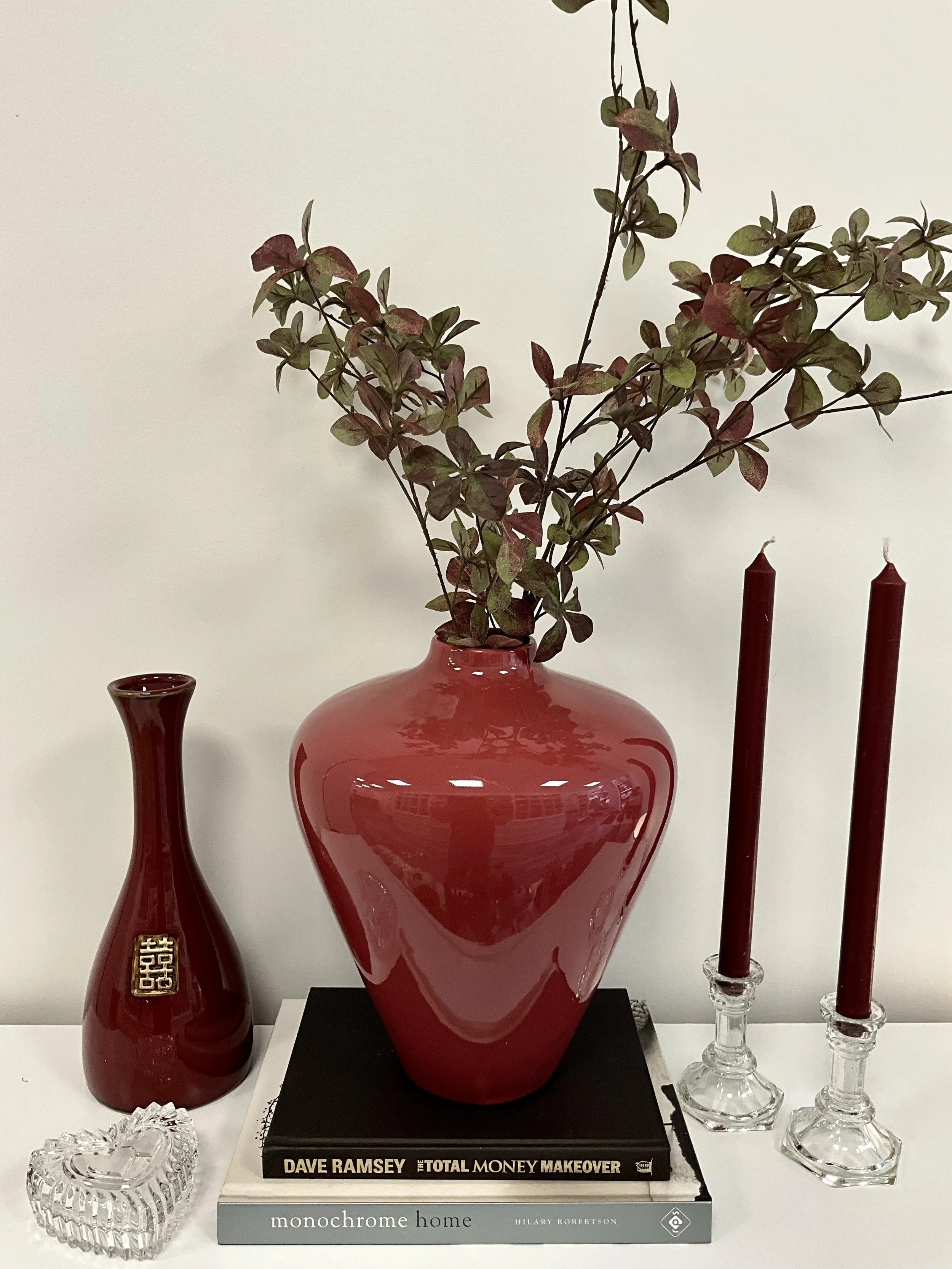 A still life arrangement featuring a large red vase with leafy branches, a smaller red ceramic vase, a pair of red candles in glass holders, and two books titled 'The Total Money Makeover' by Dave Ramsey and 'Monochrome Home' by Hilary Robertson, all
