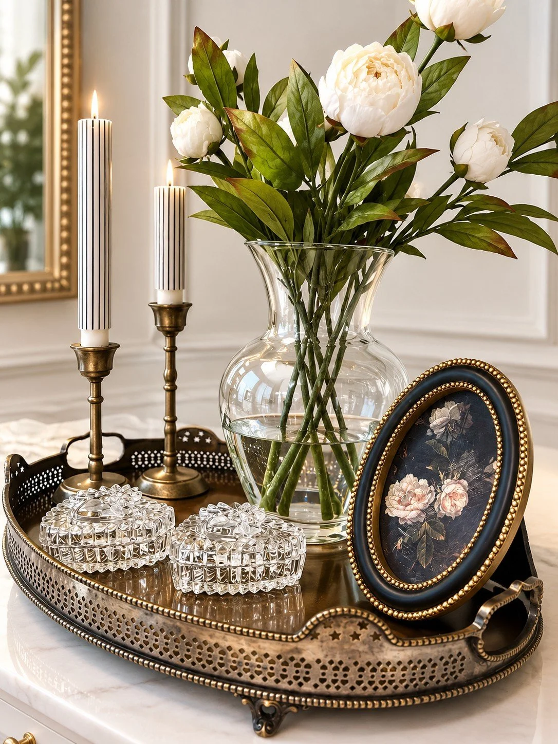 A decorative tray with a glass vase of white peonies, two tall striped candles, a framed floral picture, and small glass jewelry boxes, all arranged on a marble surface.