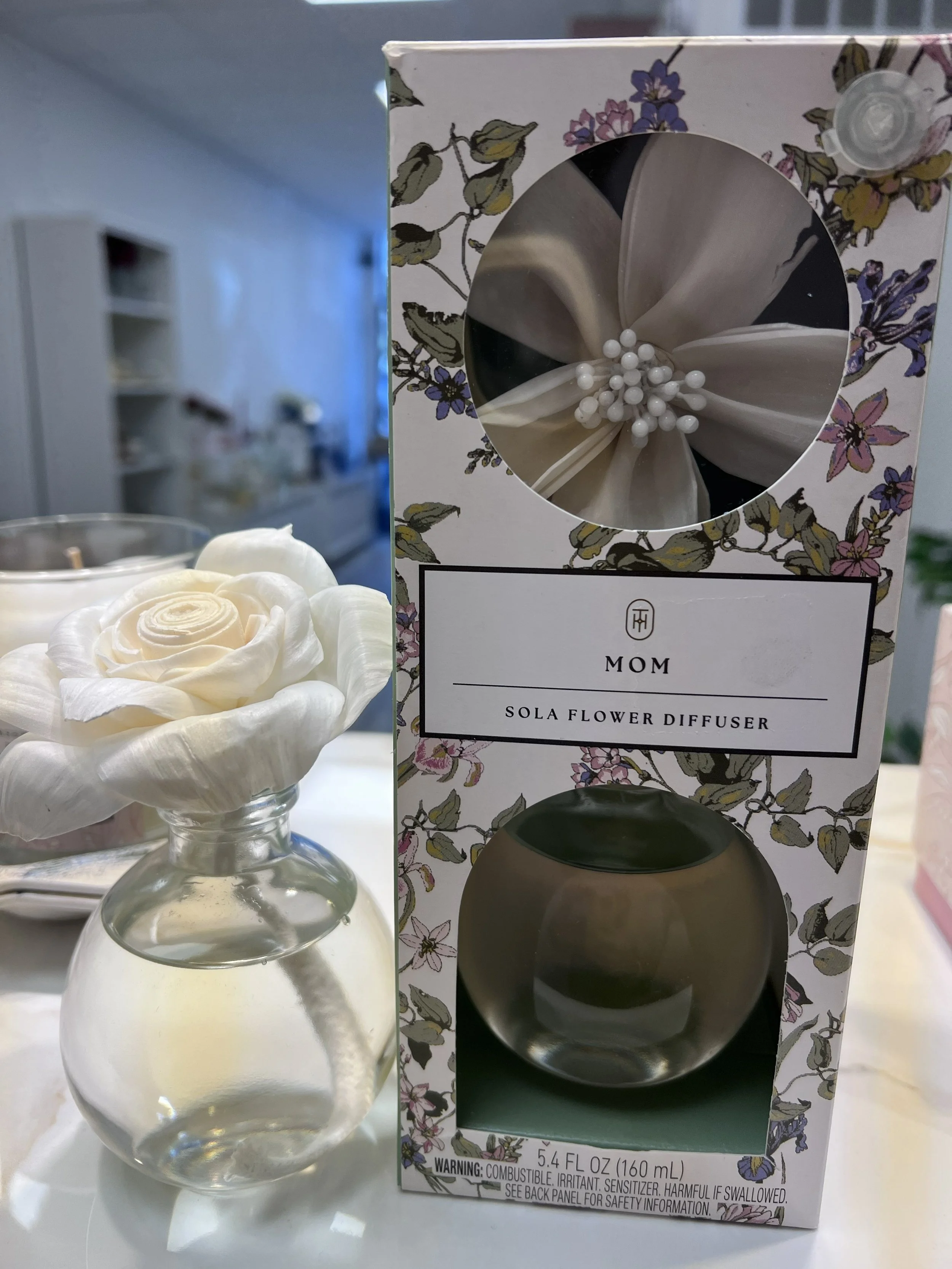 A small glass bottle with a white artificial flower arrangement next to it, and a floral patterned box containing a Sola flower diffuser in the background.