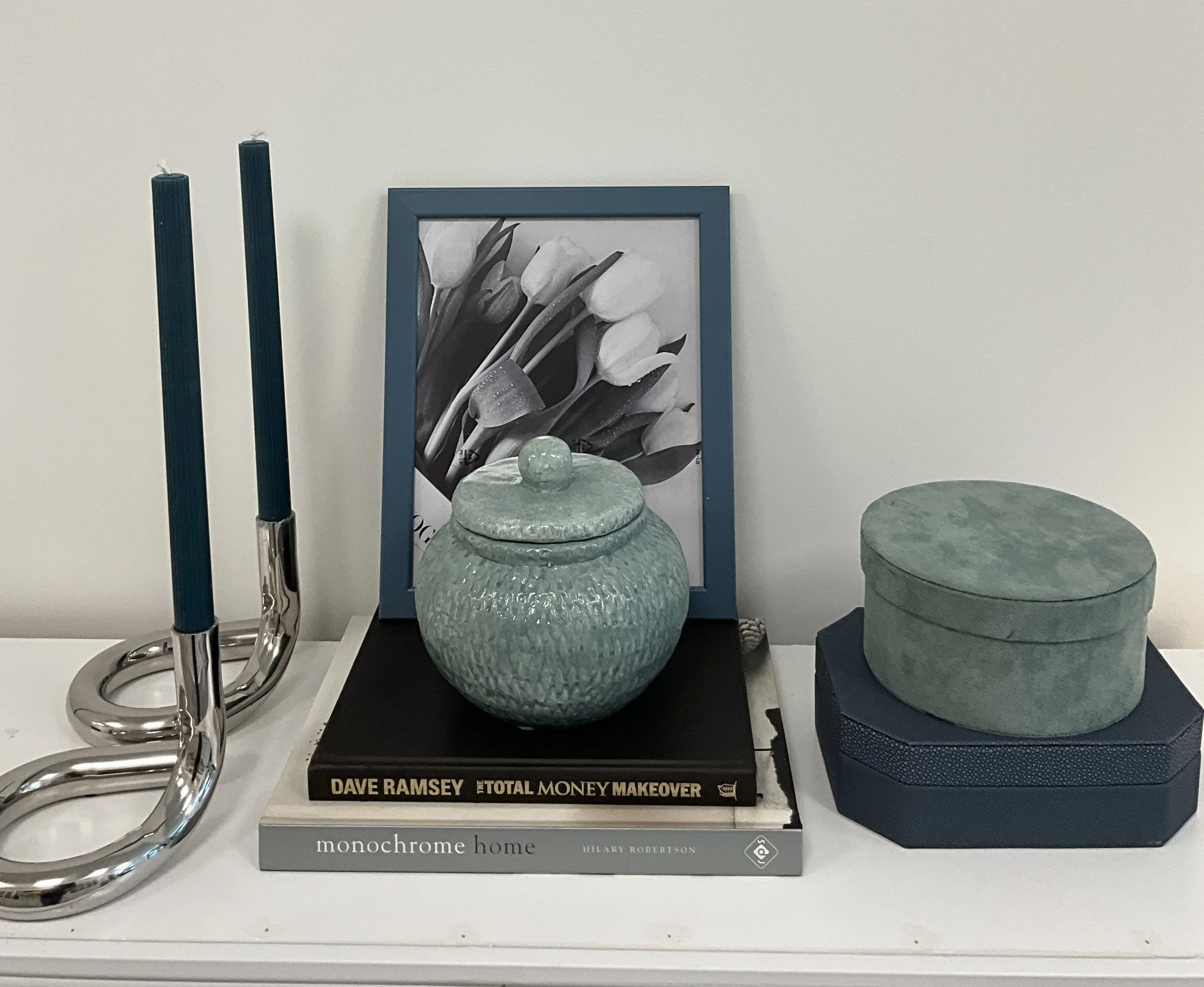 Decorative displayed items including blue candles, a framed black and white photo of white tulips, a large green ceramic jar with a lid, a book titled 'The Total Money Makeover' by Dave Ramsey, and a gray hardcover book titled 'Monochrome Home' by Hi