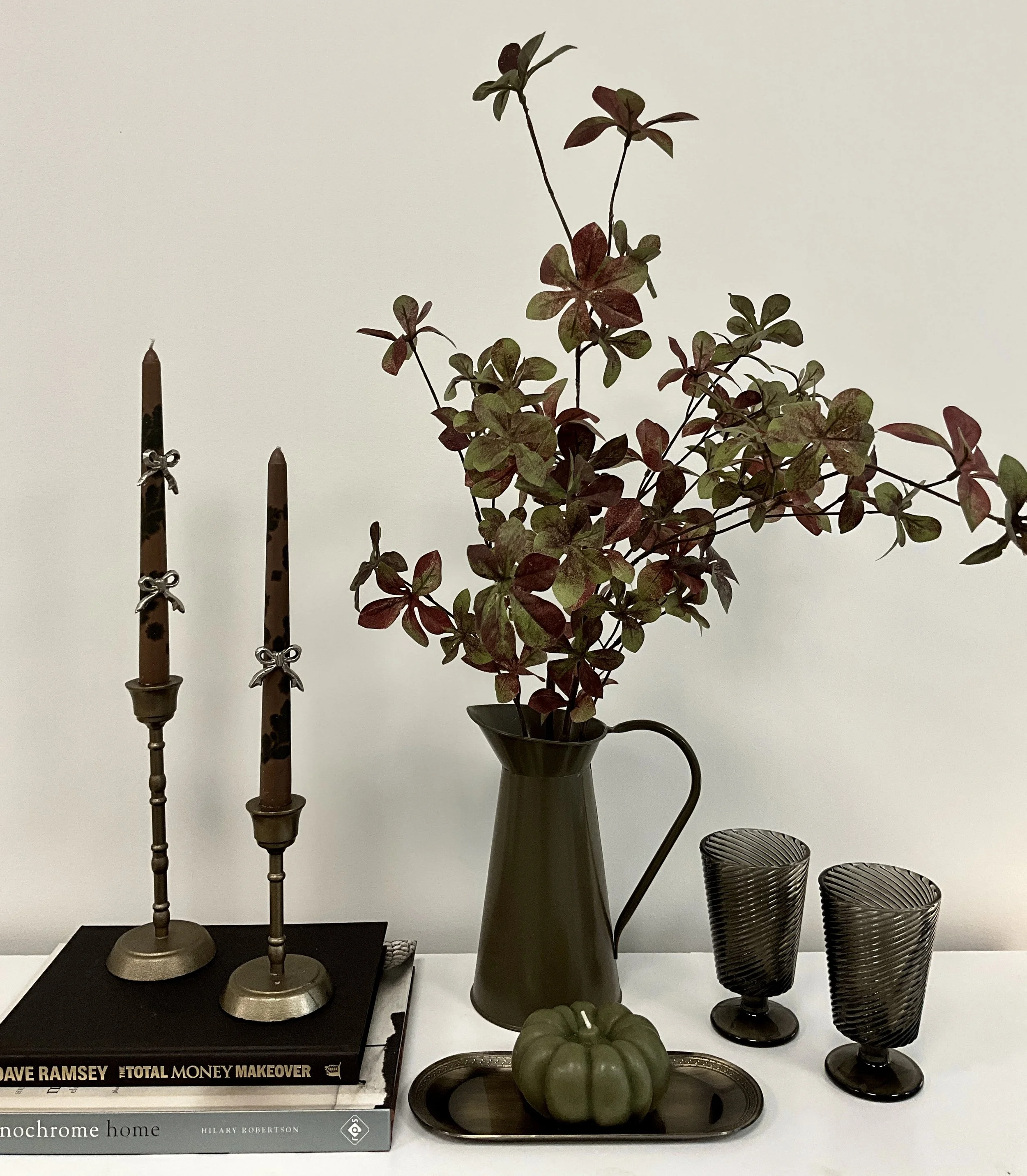 Decorative home display featuring three tall candle holders with dark and tortoise shell patterns, a black vase with a green and burgundy leafy plant, a gray decorative pumpkin, a silver tray, and three black wire baskets on a white surface against a