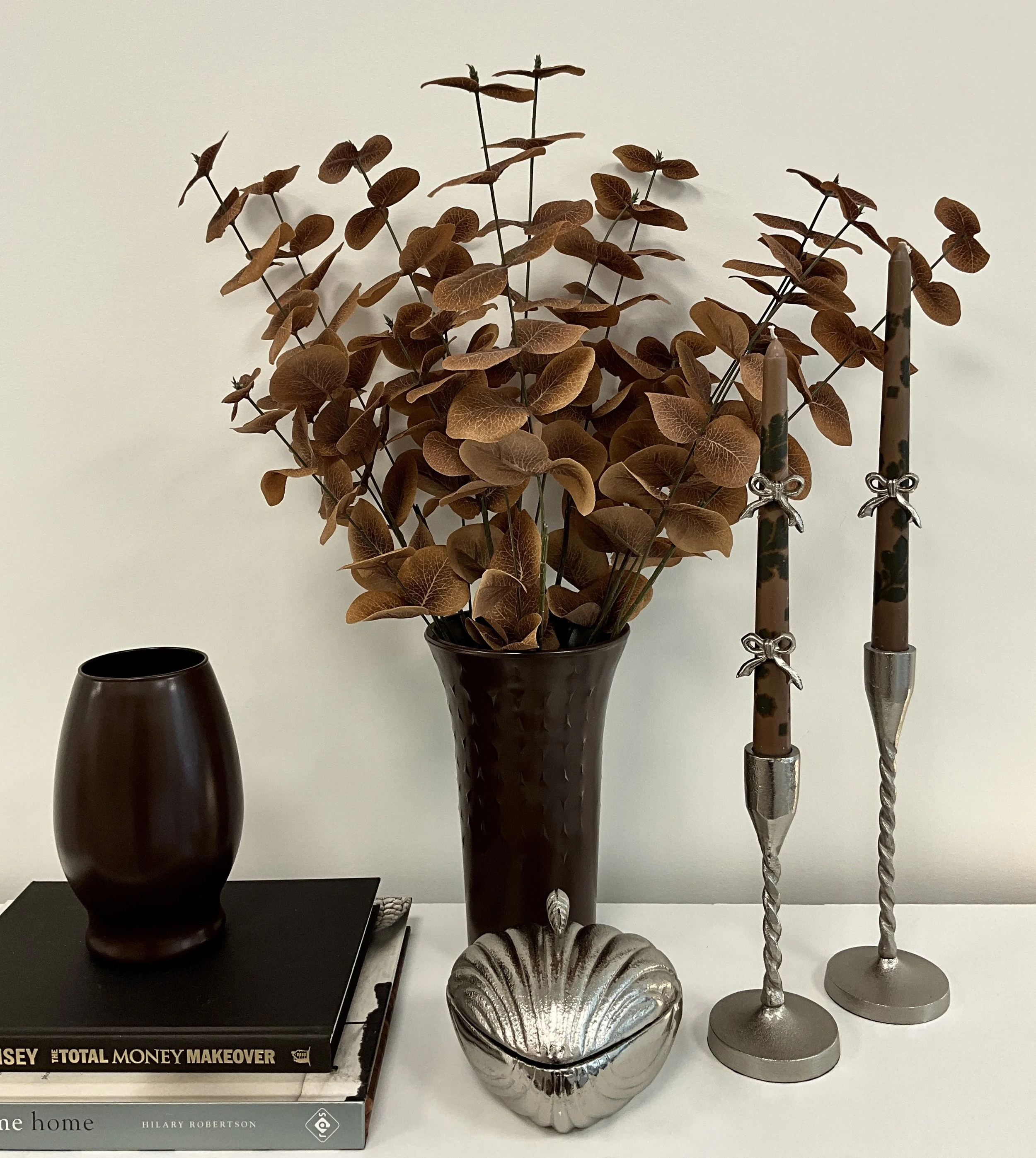 Decorative objects on a white surface, including a plant with brown leaves in a dark brown vase, three candlesticks with unlit candles, and two stacked books with black and white covers.