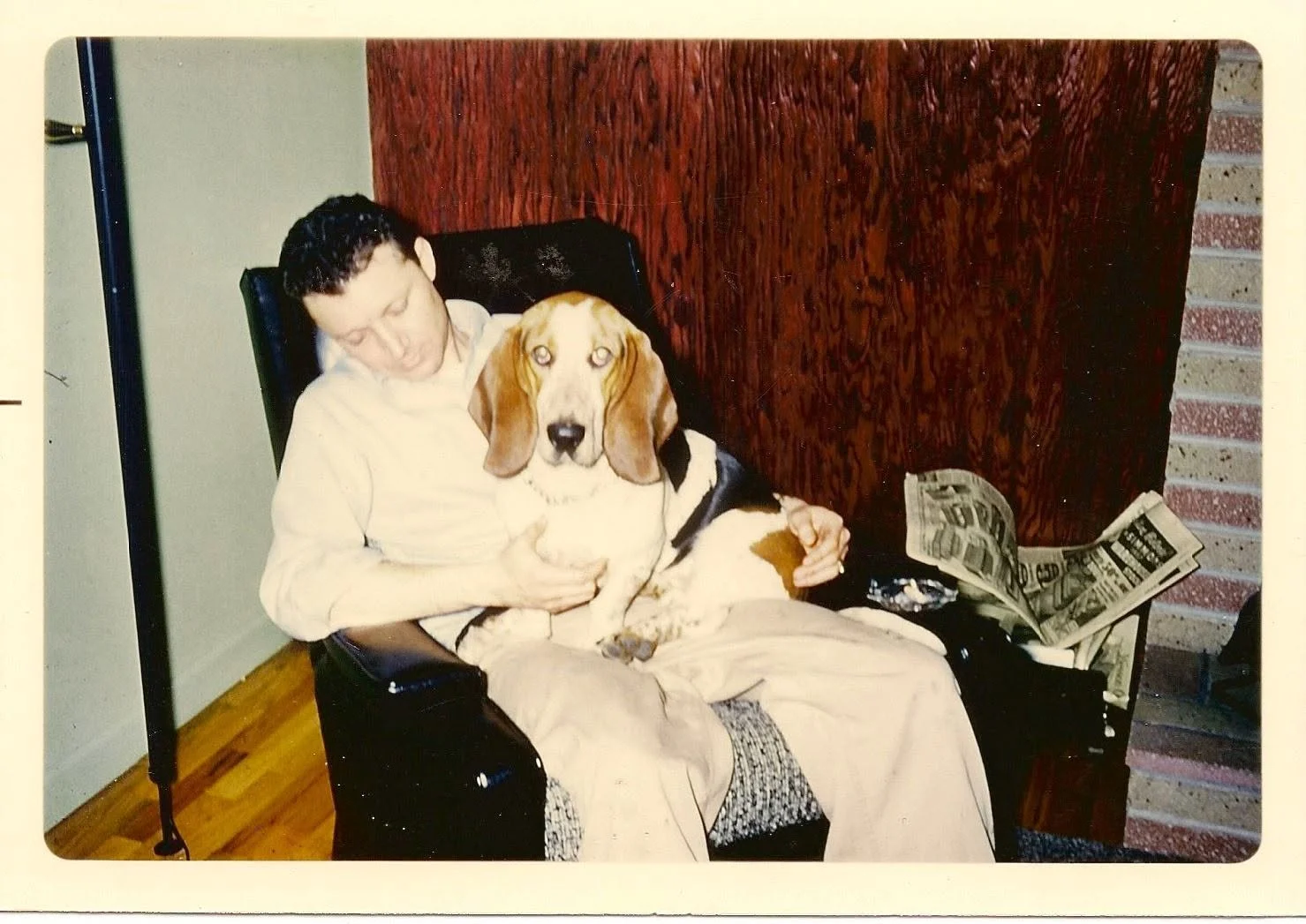 A man sitting in a chair holding a large Beagle dog on his lap, with a newspaper and some dollar bills resting on the armrest.