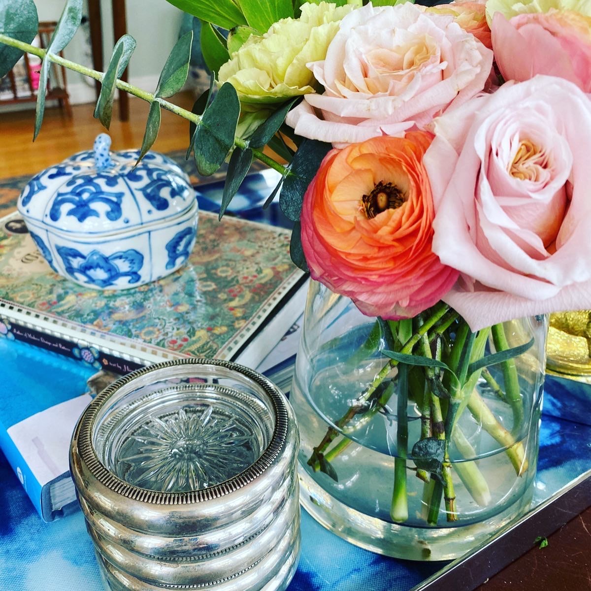 A bouquet of pink, peach, and cream roses with green leaves in a clear glass vase sits on a table. Nearby is a stack of small, round glass dishes, and a blue and white patterned ceramic jar with a lid. In the background, there is a table with a decorative box or book with a colorful cover.