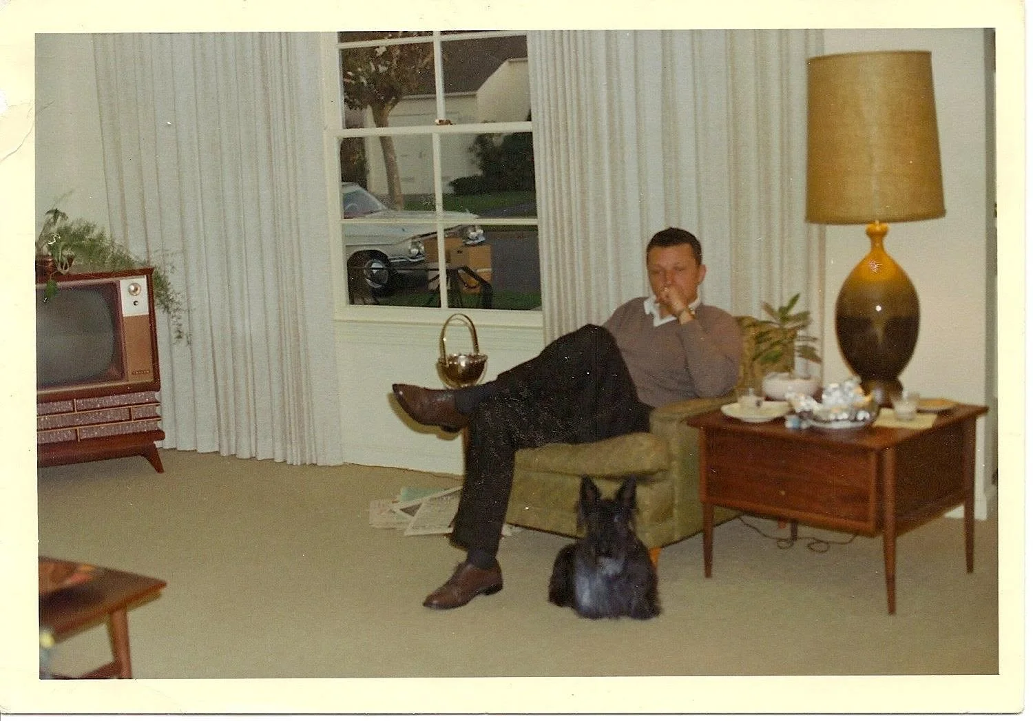 A vintage photo of a man sitting in a living room. He is sitting on a green armchair, with one leg crossed over the other. There is a black dog sitting on the floor in front of him. The room has beige carpet, a wooden side table with a large lamp, a window with white curtains, and an old television set.