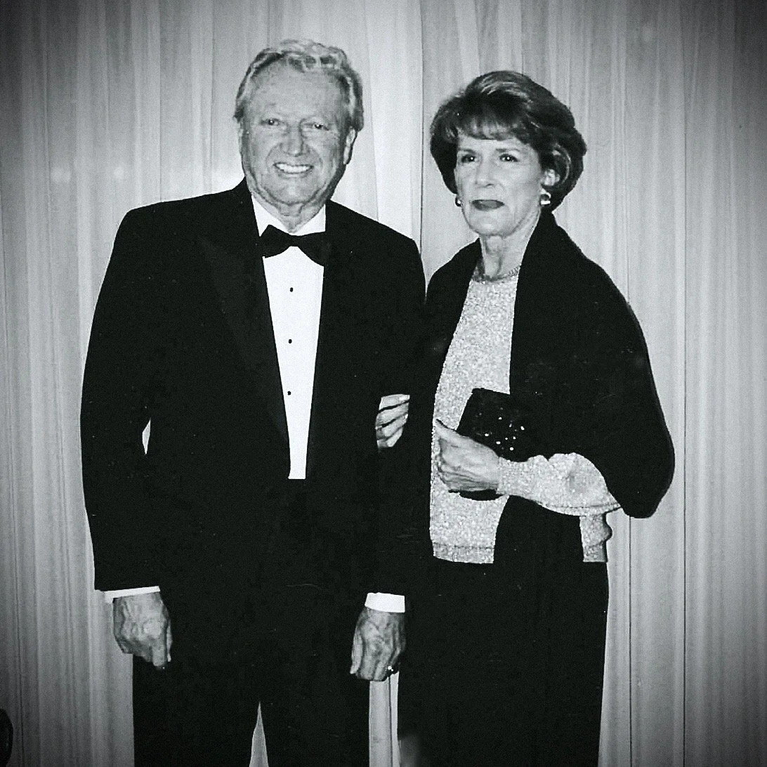 Black and white photo of a man in a tuxedo and a woman in an elegant outfit standing together against a wooden panel background at a formal event.