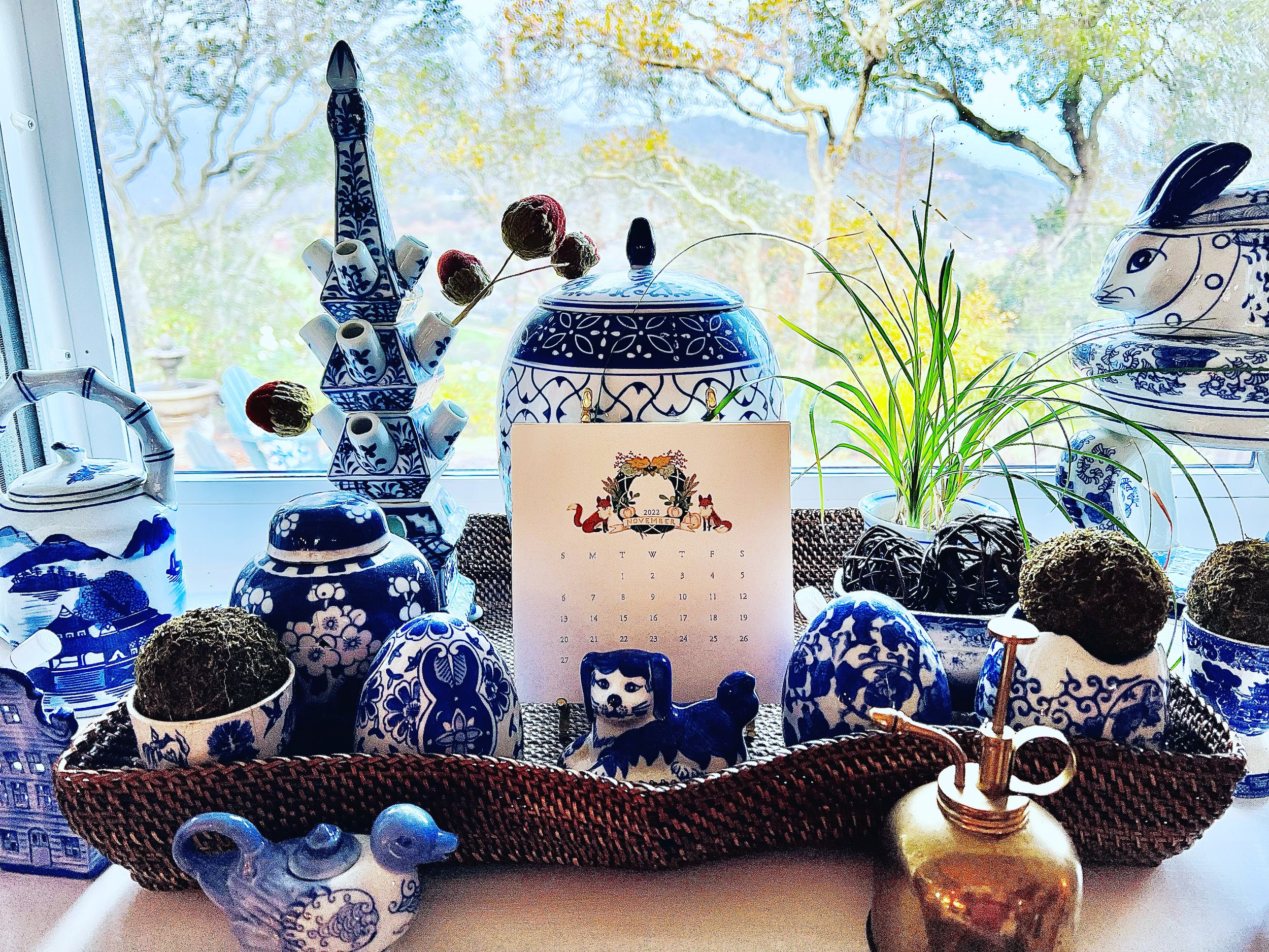 A collection of blue and white ceramic vases, bowls, and figurines with floral and animal patterns, arranged on a surface near a window with a view of trees outside, and a calendar showing November 2022 at the center.