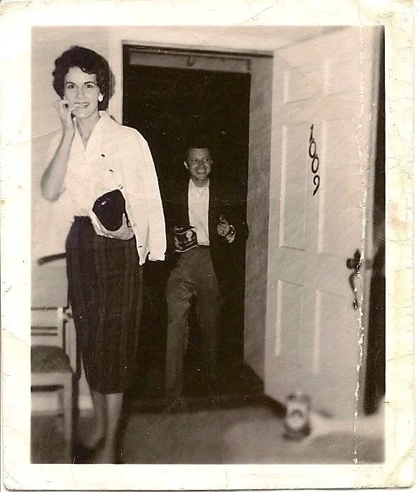 A black-and-white photo of a woman and a boy in a house. The woman is standing outside a room, smiling, with her hand on her face, wearing a white blouse and a dark skirt. The boy is in the doorway, also smiling, holding a camera in each hand, wearing a dark jacket and pants.