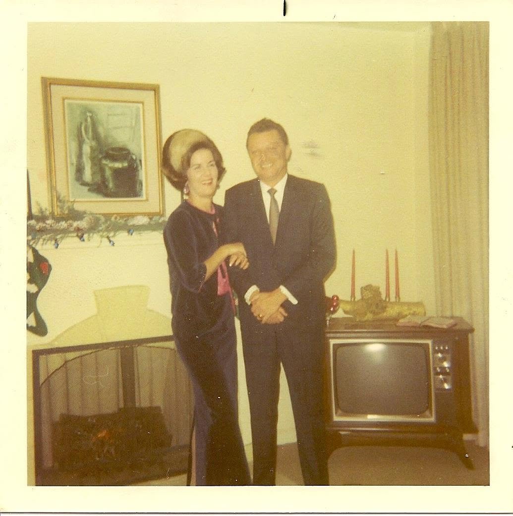 A vintage photograph of a woman and a man dressed in formal attire, standing together in a living room decorated for Christmas with a fireplace, artwork, and a television.