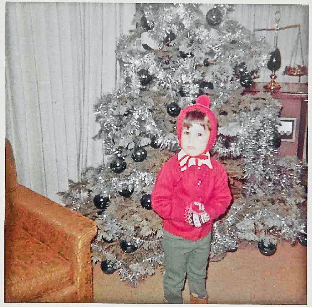 A young boy dressed in a red coat, knit hat, and gloves standing in front of a decorated Christmas tree in a living room.