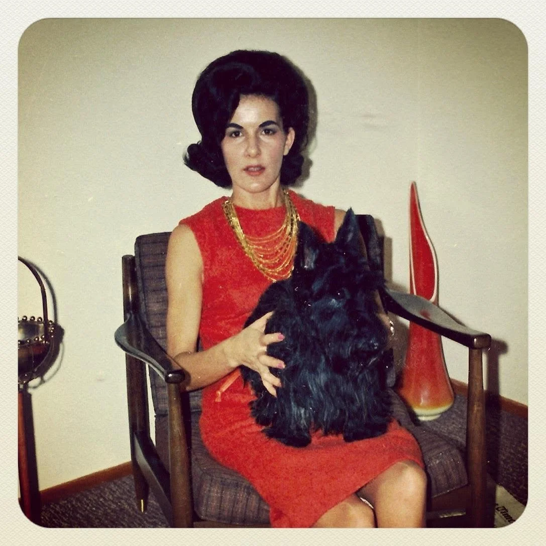 Woman with dark, voluminous hair sitting on a wooden chair, wearing a red dress and gold jewelry, holding a black dog.
