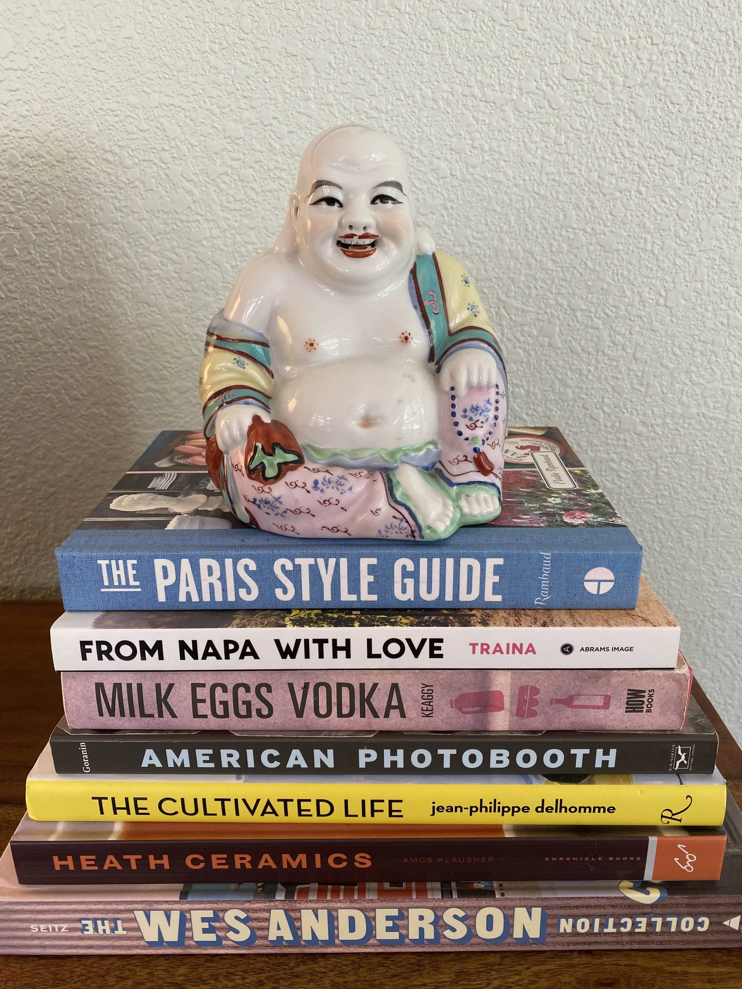Stack of six books on a wooden surface. The top book is titled 'The Paris Style Guide' and features a ceramic laughing Buddha figurine with bright colors and detailed patterns on top. The books below cover topics like wine, vodka, photography, life, ceramics, and city writing, with visible titles including 'From Napa With Love,' 'Milk Eggs Vodka,' 'American Photo Booth,' 'The Cultivated Life,' 'Heath Ceramics,' and 'The West Anderson'.