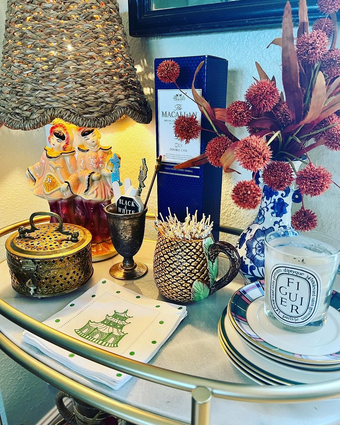Decorative table with a blue box of whisky, a vase with pink flowers, a ceramic pineapple-shaped pitcher, a candle holder, a small black container with toothpicks, and a candle labeled 'Figuer' with a plate underneath. There are also napkins with a green pagoda design placed on the table.